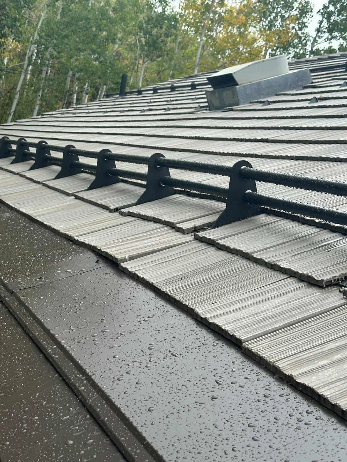 Close-up of a roof with gray shingles, black metal brackets, and a vent, with wet surface and droplets of rain, and trees in the background.