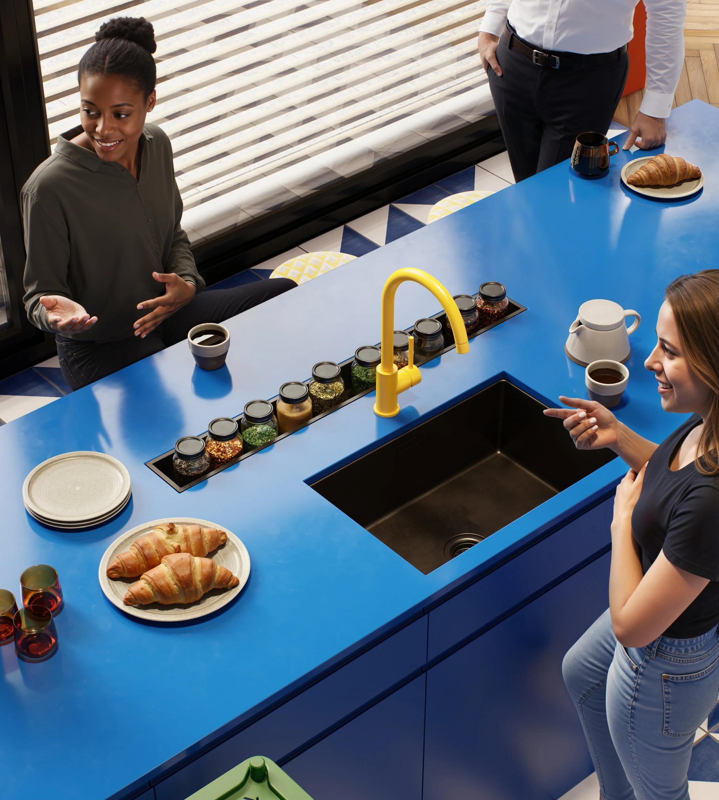 Two women and a man in a kitchen with a bright blue countertop. The women are talking, and the man is standing nearby. There are two croissants on a plate, jars of spices, cups of coffee, and a teapot on the counter. A large window with blinds is in 