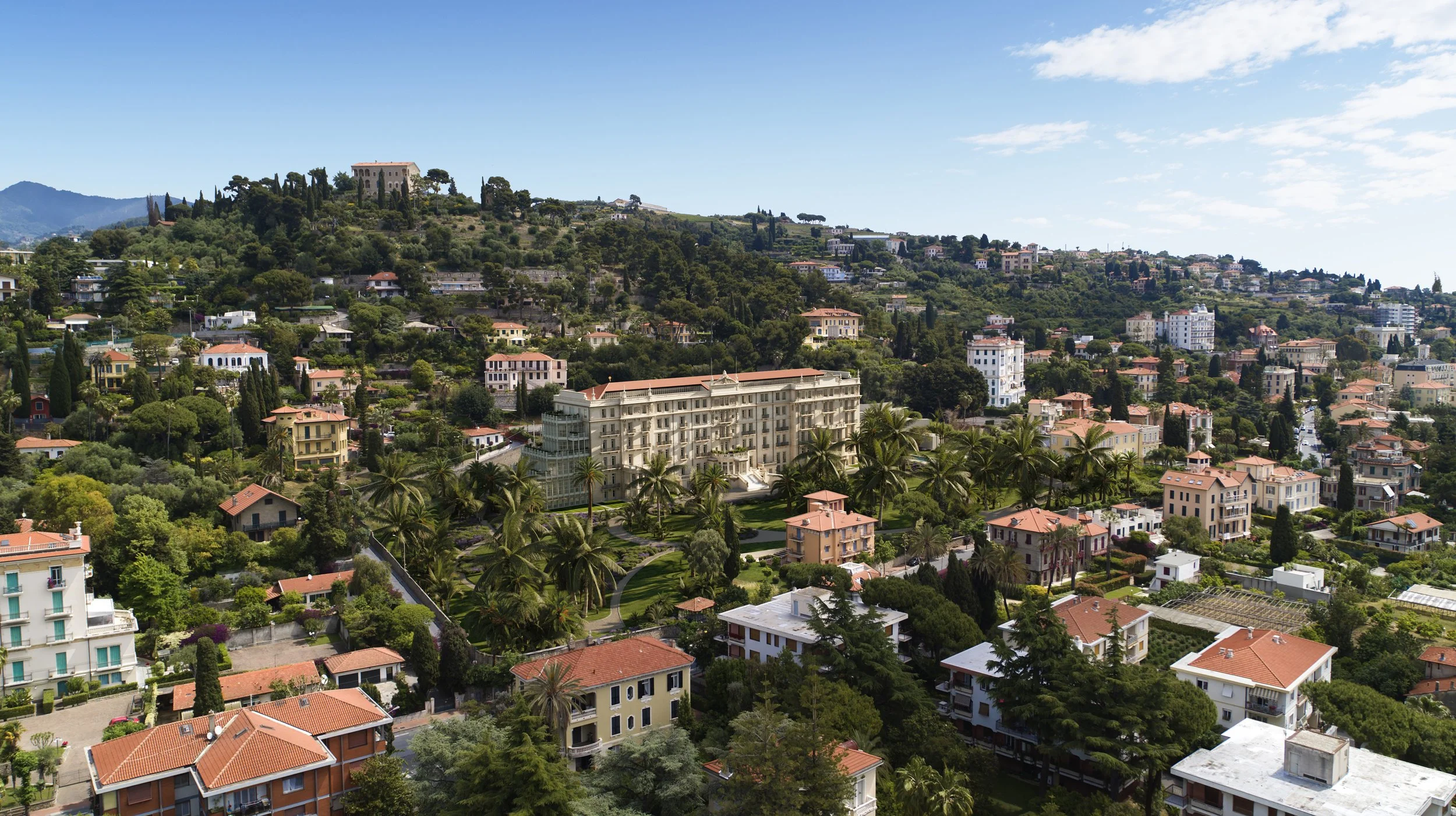 Aerial view of a hillside with numerous houses and greenery, featuring a large white hotel with palm trees in the foreground, under a partly cloudy blue sky.