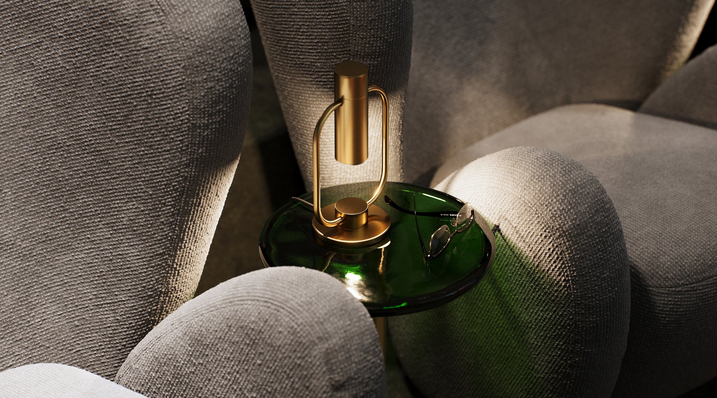 A small modern lamp with a gold finish on a round green glass table between two gray upholstered armchairs. A pair of glasses rests on the table.