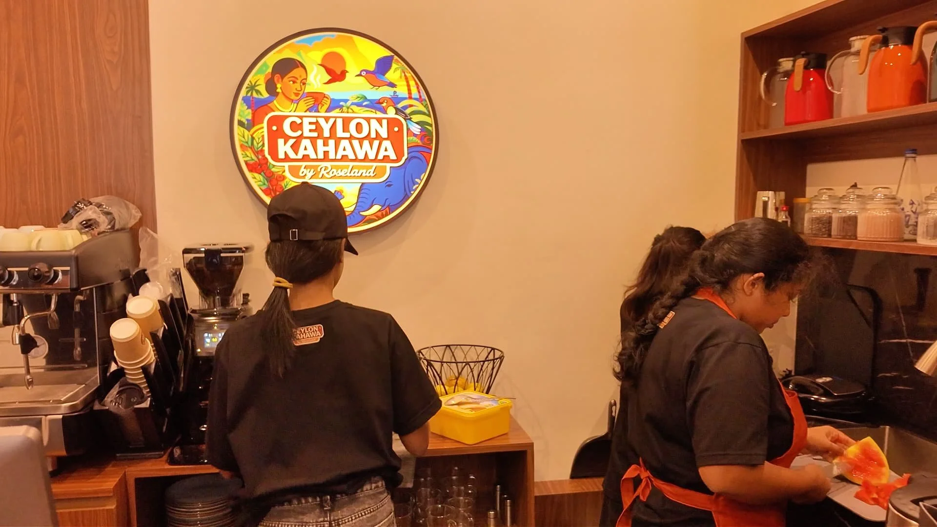 Roseland Coffee Roasters' first cafe, Ceylon Kahawa, with beautiful colourful logo depicting a woman, sunrise, birds, coffee plant and cherries, hills and an elephant. Three employees are working behind the counter and making coffee, facing the logo.