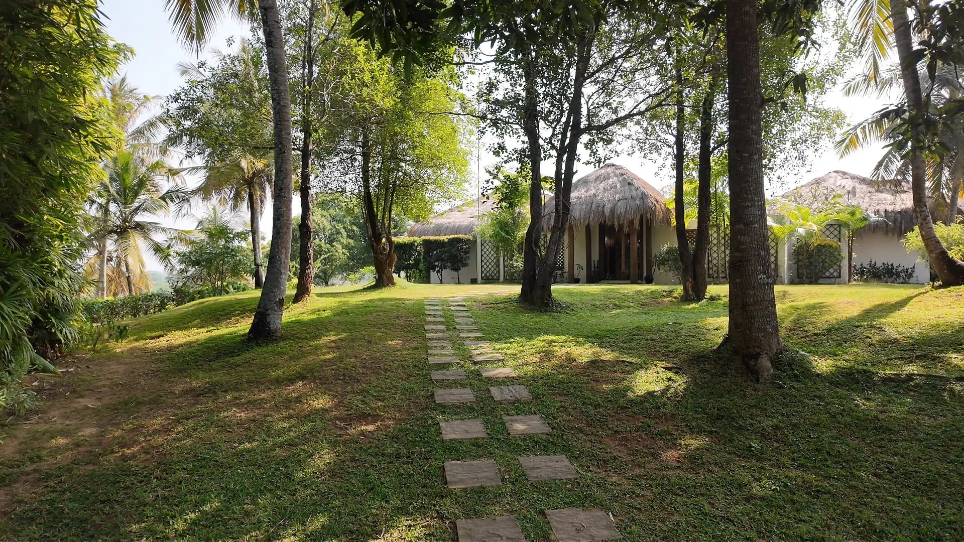 A tropical garden path leading to a thatched-roof cottage of Tekanda Lodge in south Sri Lanka surrounded by trees and lush greenery.
