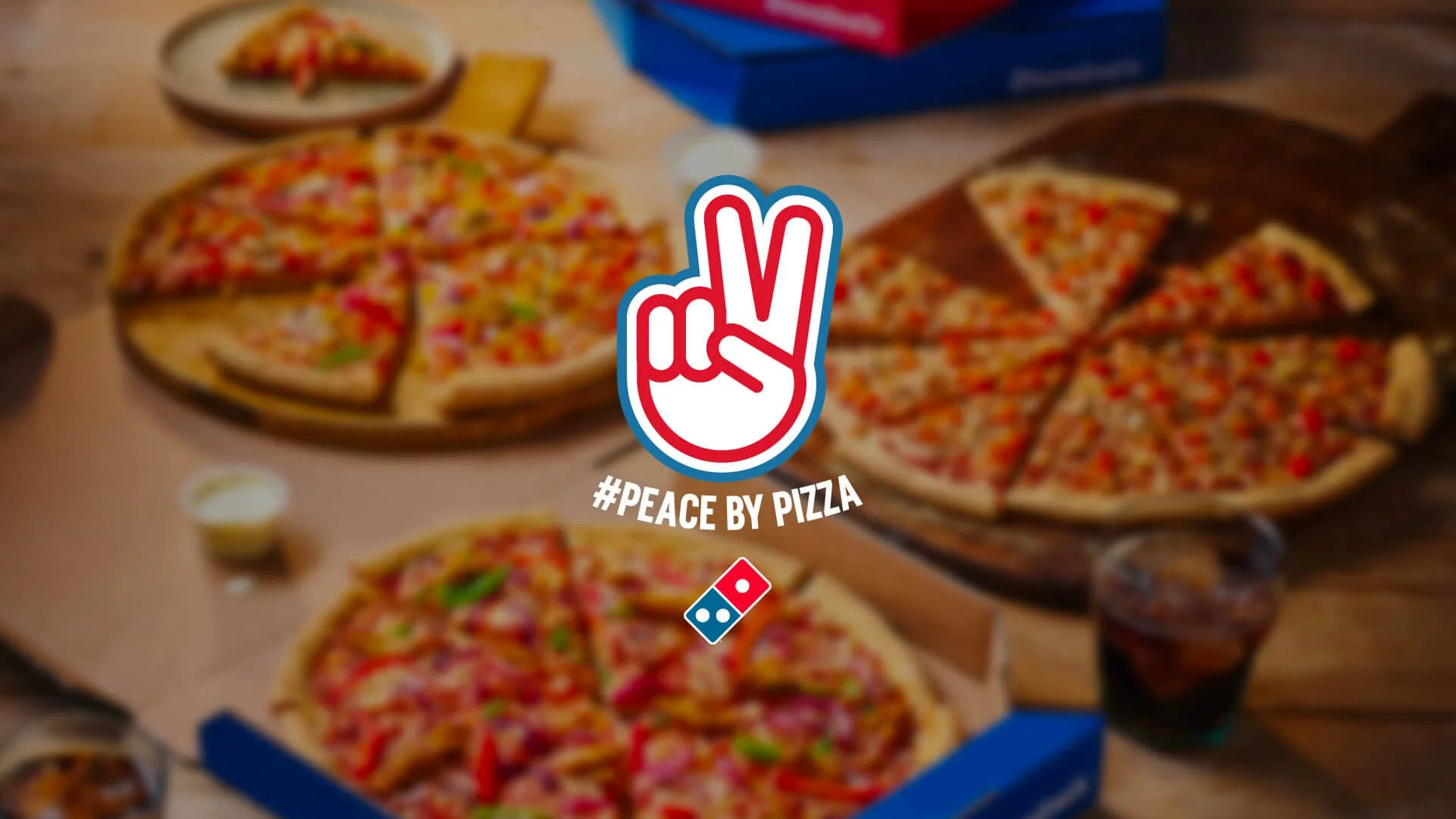 Three pizzas on a table with a Peace by Pizza sticker and a Domino's logo in the center. The pizzas are topped with various ingredients and are cut into slices. There are drinks and dipping sauces around the pizzas.