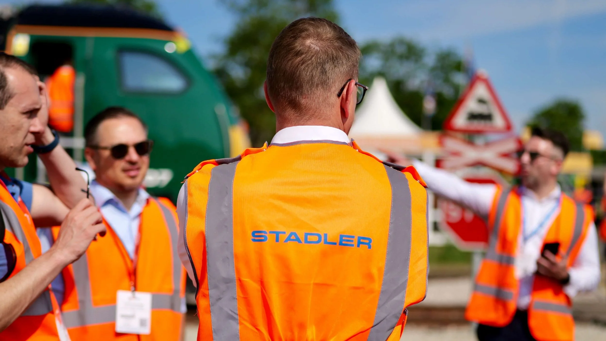 Group of people wearing bright orange safety vests, standing outdoors at a construction or event site, with one person facing away from the camera and others talking nearby.