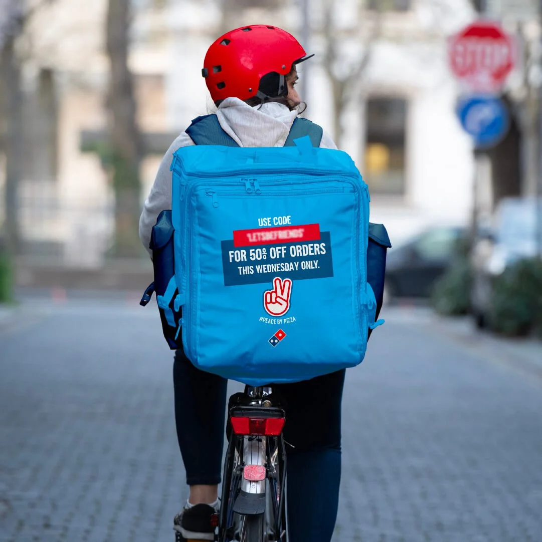 Person riding a bicycle wearing a red helmet and backpack with a pizza delivery promotion, on a city street.
