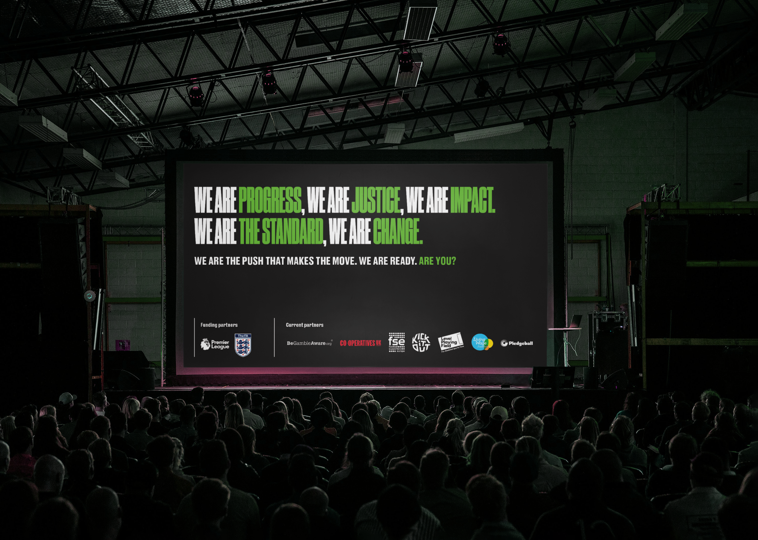 Large screen on stage displaying a motivational message with green and white text, surrounded by an audience in a dark indoor venue, with stage lighting and equipment overhead.