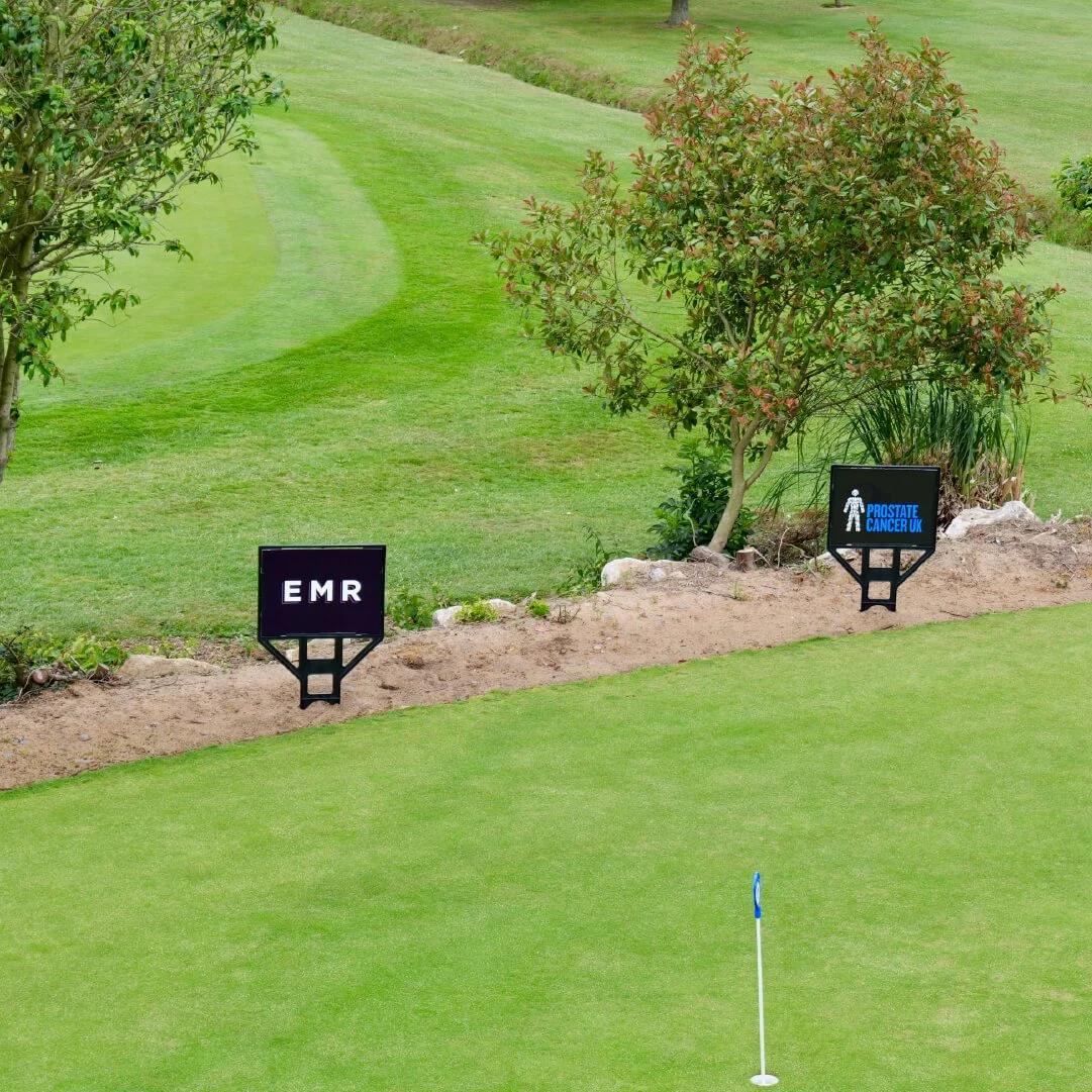 A golf course green with a blue flag, two signs on the edge reading EMR and PROSTATE CANCER UK, and trees and bushes in the background.