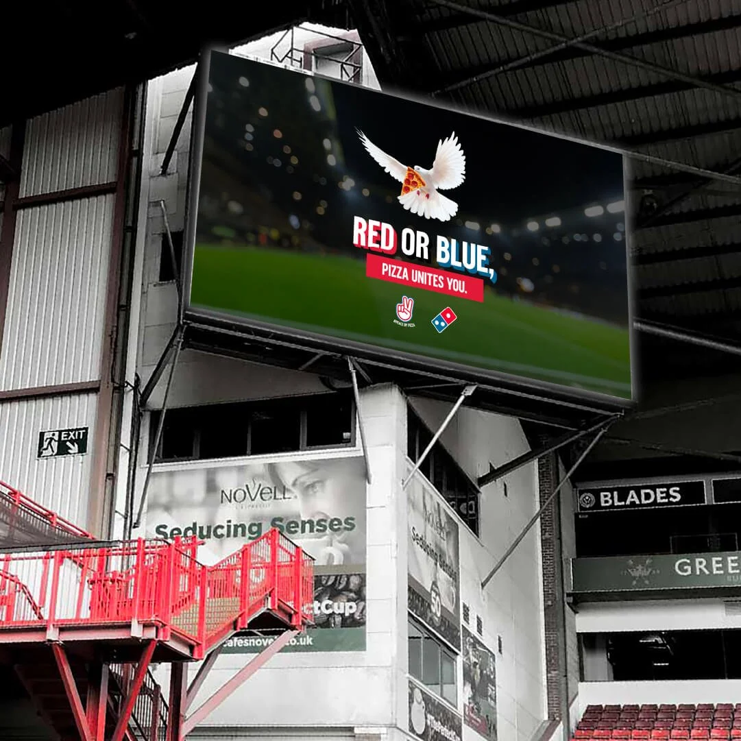 A digital billboard inside a stadium features an ad with a dove holding a piece of pizza, promoting a pizza chain with the slogan 'Red or Blue, Pizza Unites You,' set against a blurred stadium background.