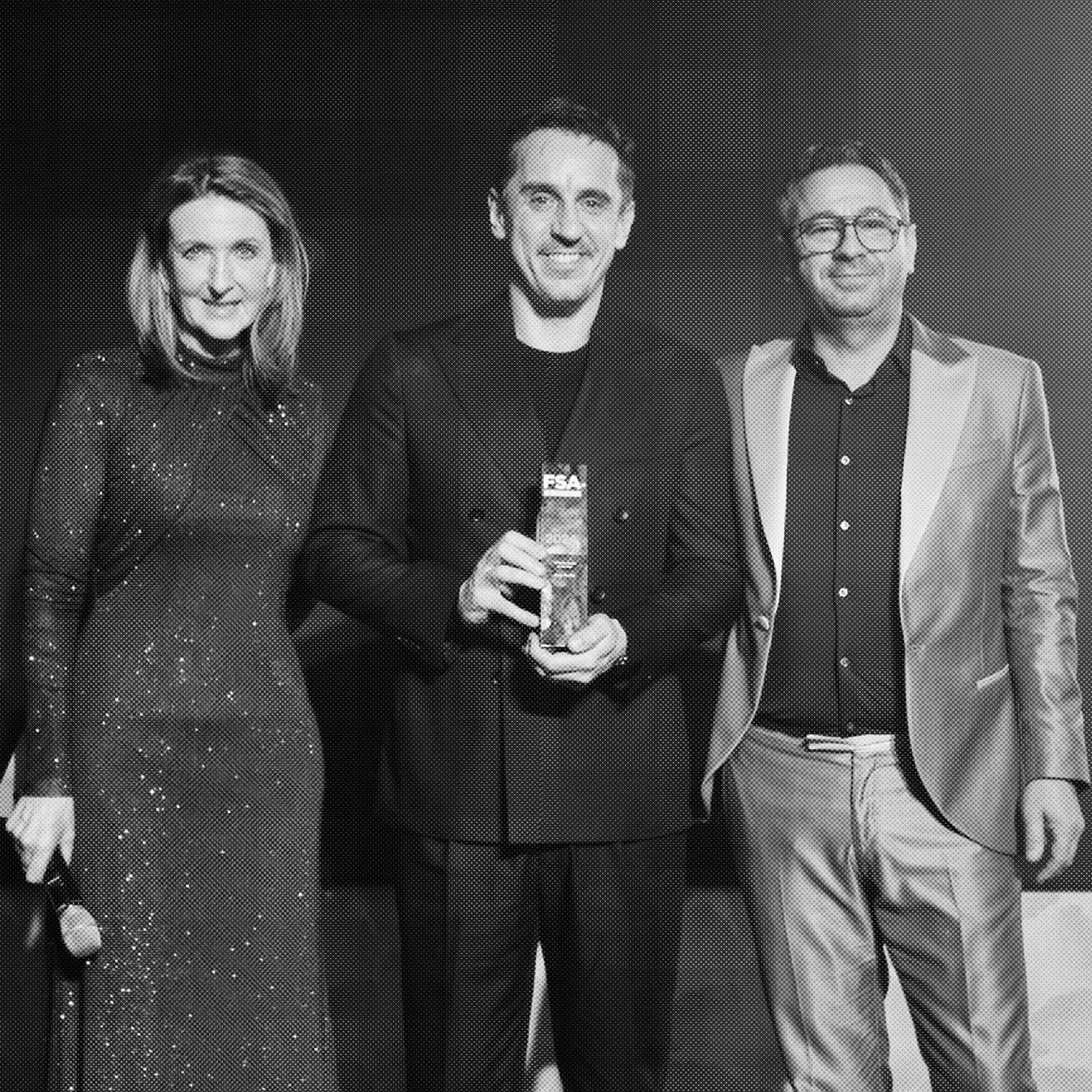 Three people standing together, one man in the middle holding a glass and an award, woman on the left wearing a sparkly dress, man on the right wearing a suit with a satin lapel.