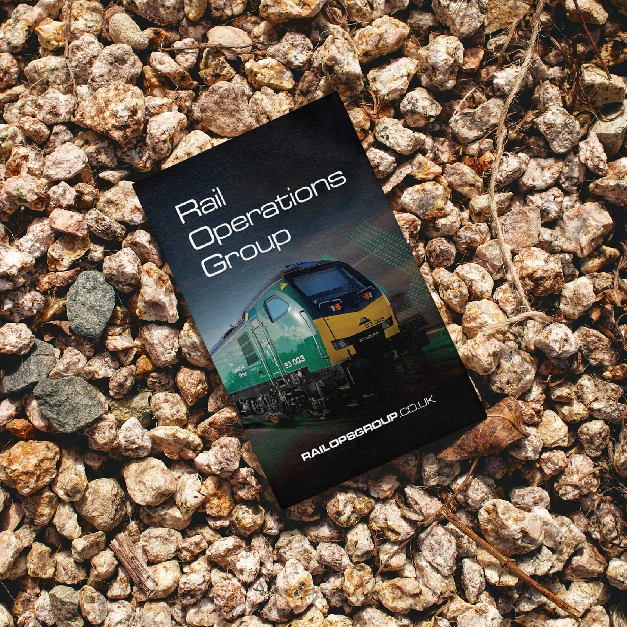 A brochure for the Rail Operations Group lies on gravel. The brochure features an image of a green and yellow train and the website URL railopsgroup.co.uk.