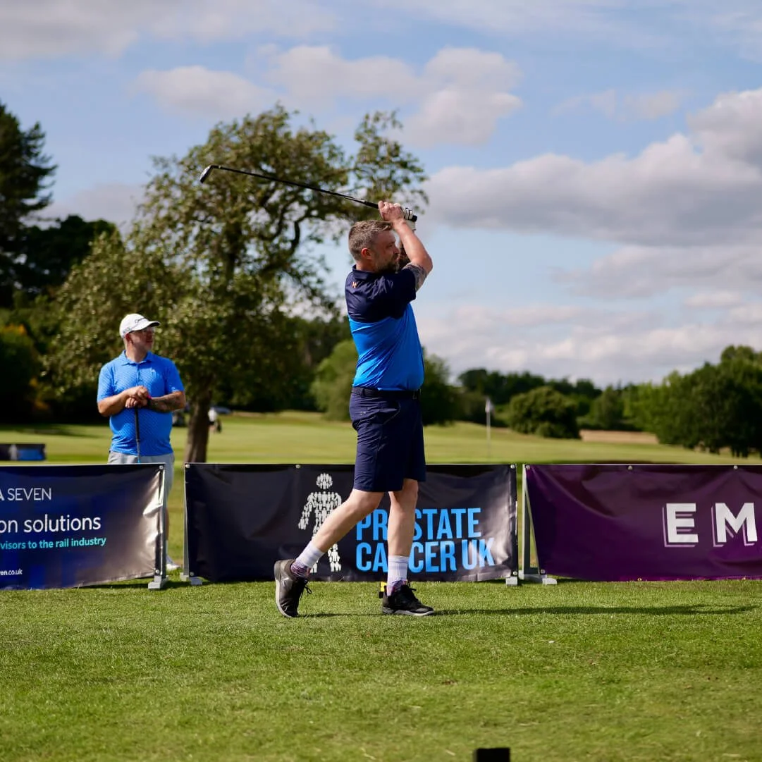 A man swinging a golf club on a golf course with another man nearby, and advertising banners in the background.