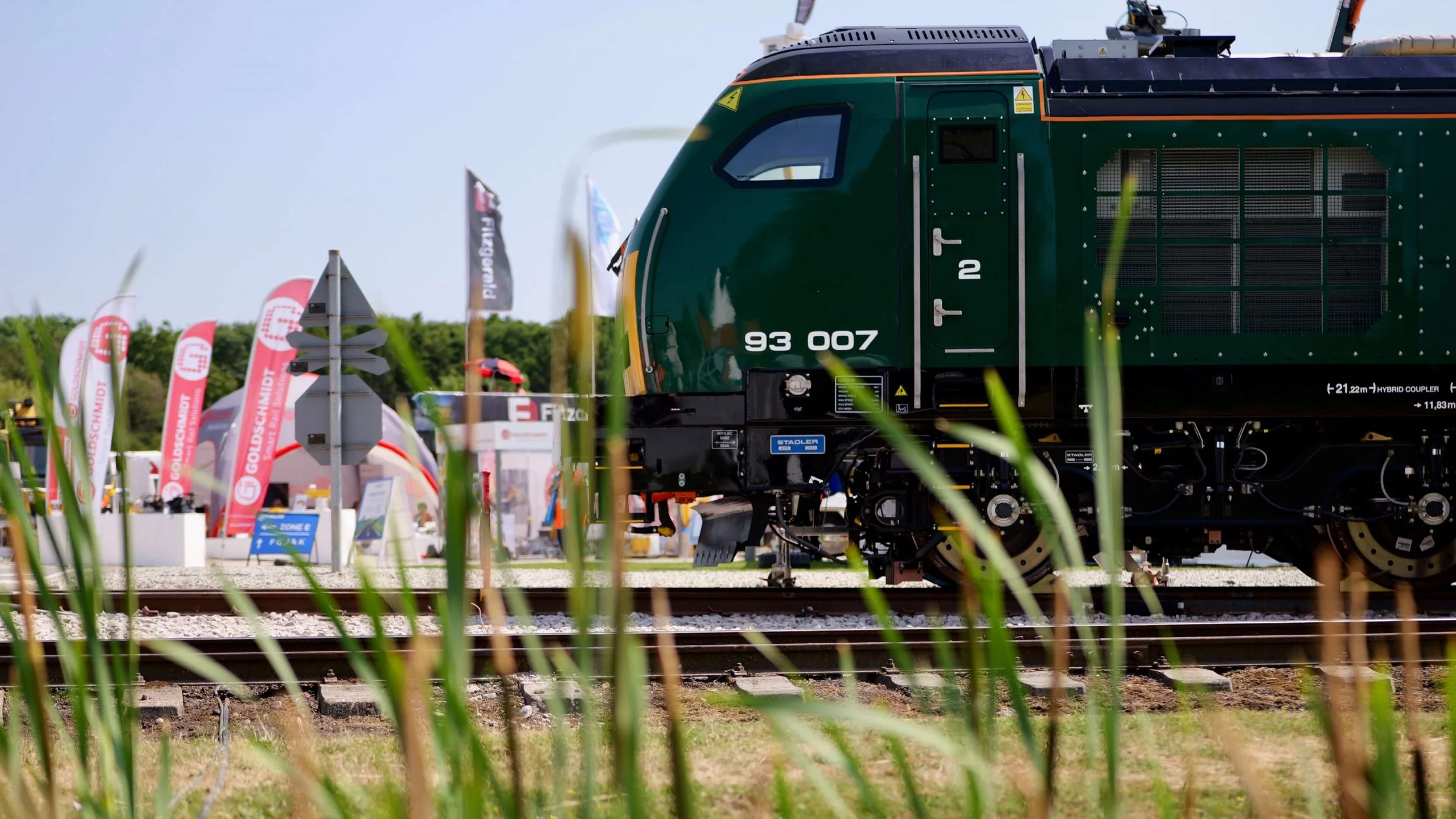 A green train locomotive at an outdoor exhibition with signs and flags in the background, partially obscured by tall grass in the foreground.