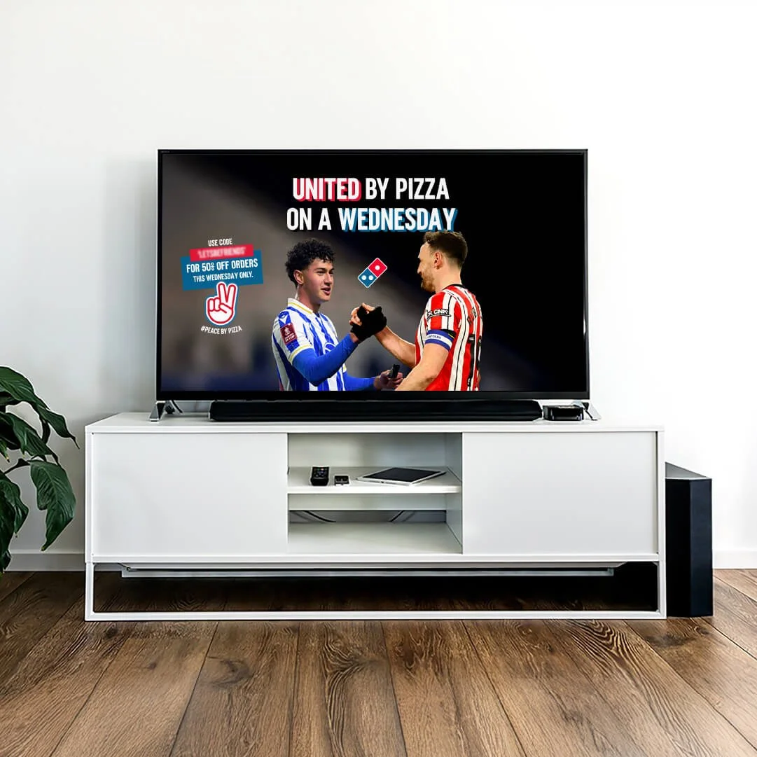 Smart TV on a white stand displaying an advertisement for Domino's Pizza, featuring two soccer players shaking hands, with text promoting a Wednesday pizza offer.