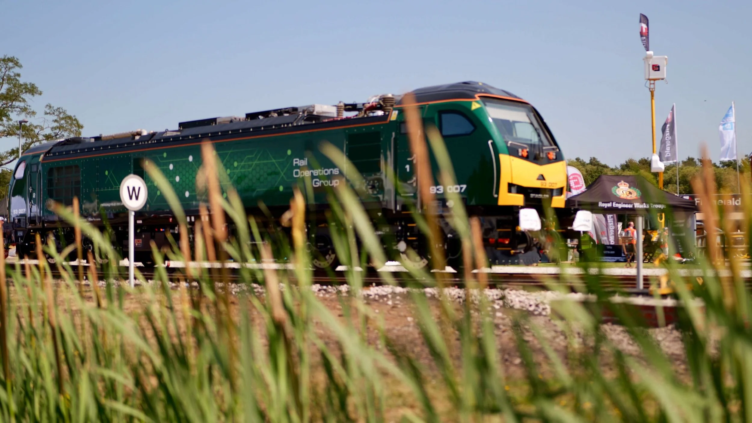 Green and black train with yellow front, labeled 'Rail Operations Group,' on a railway track outdoors with flags and a small tent nearby, and grass in the foreground.
