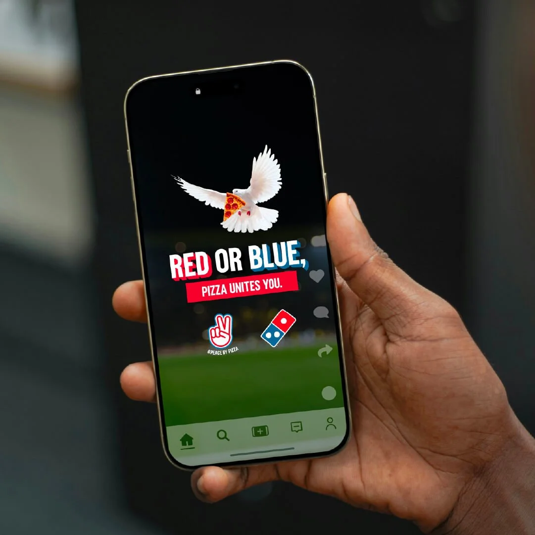 A person holding a smartphone displaying a graphic of a dove with a pizza slice on its back and the text 'Red or Blue, Pizza Unites You,' along with logos of peace and pizza.