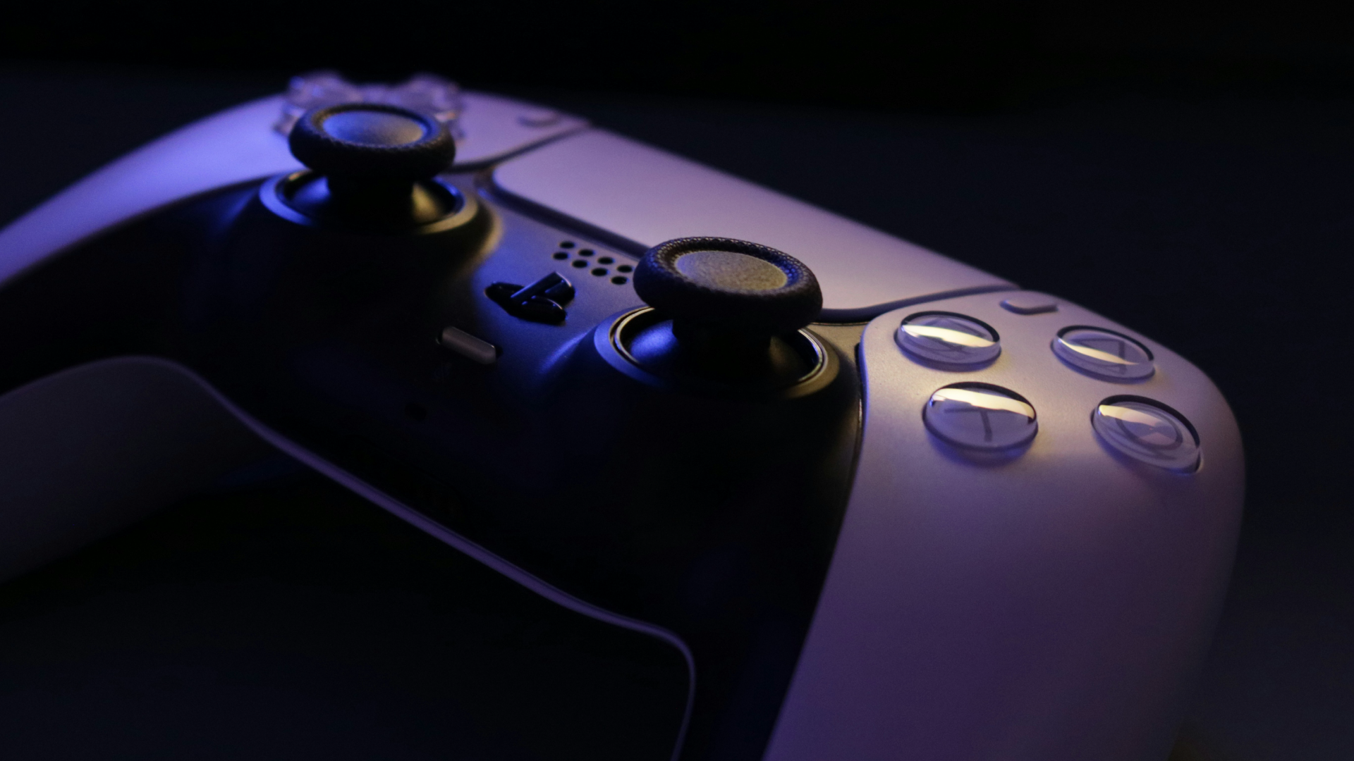 Close-up of a white gaming controller with black joysticks and buttons, illuminated by blue light in a dark setting.