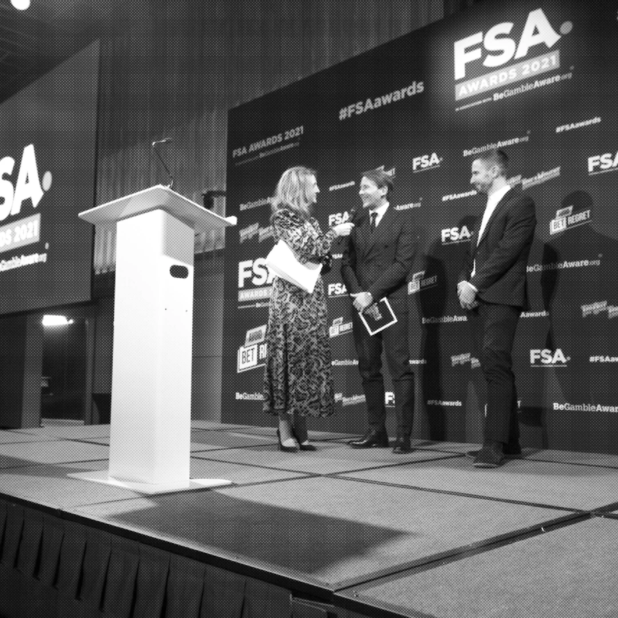 A woman in a patterned dress is interviewing a man in a suit on stage at the FSA Awards 2021. Another man in a suit stands nearby. The background features a large display with the event's logo and sponsors.