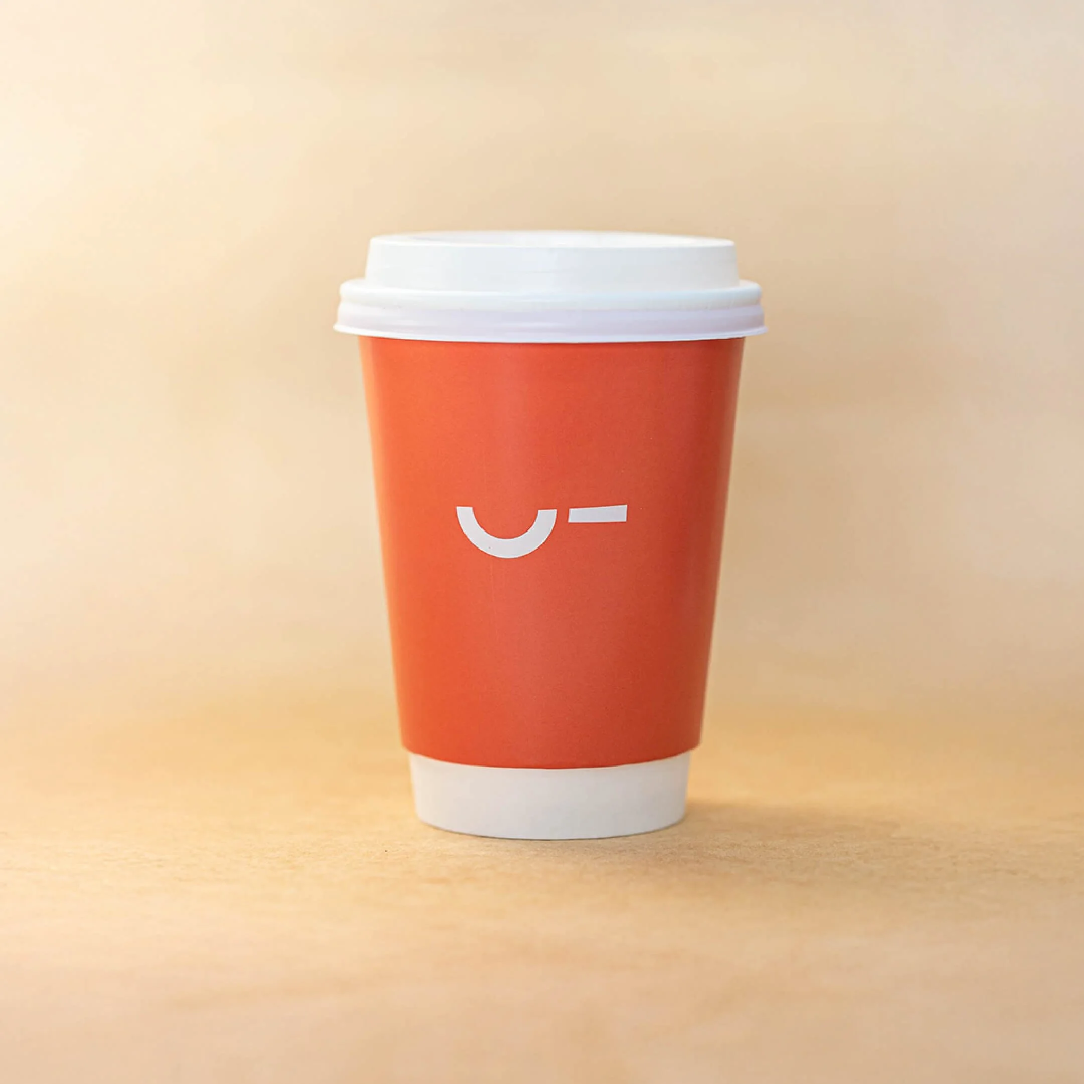 A disposable coffee cup with a white lid, orange sleeve with a white minimalist logo, placed on a light wooden surface against a beige background.