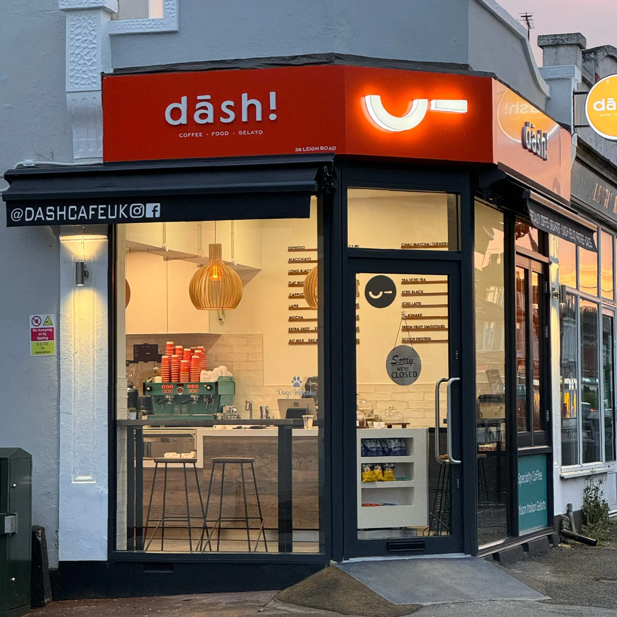 Front view of a modern coffee shop named 'dash!' with large glass windows and door, illuminated sign with a smiley face, interior with pendant lights and espresso machine, and a 'closed' sign on the door.