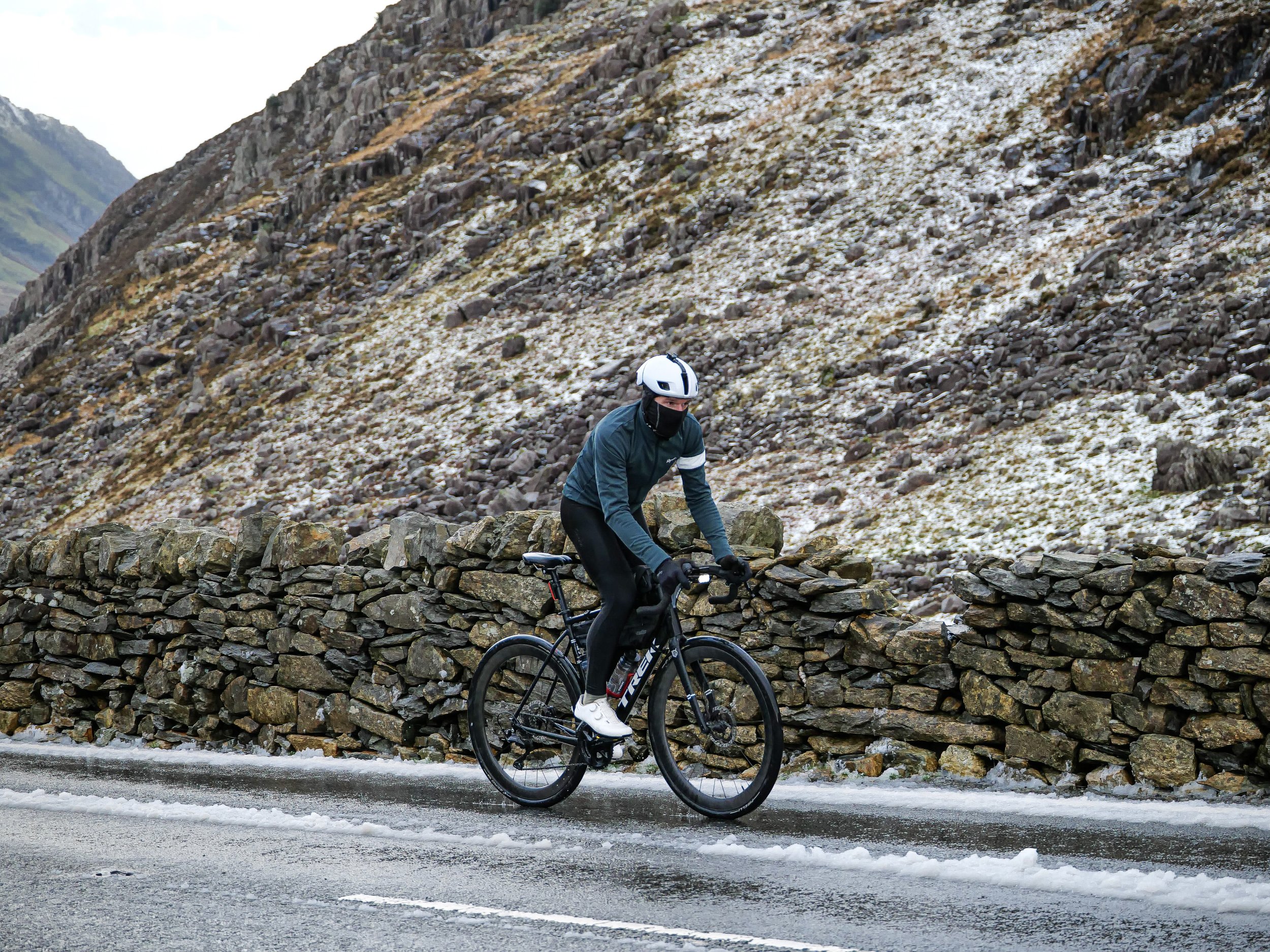 A cyclist wearing a helmet, face mask, and gloves riding a black Trek bicycle on a wet mountain road with snow, rocks, and mountains in the background.