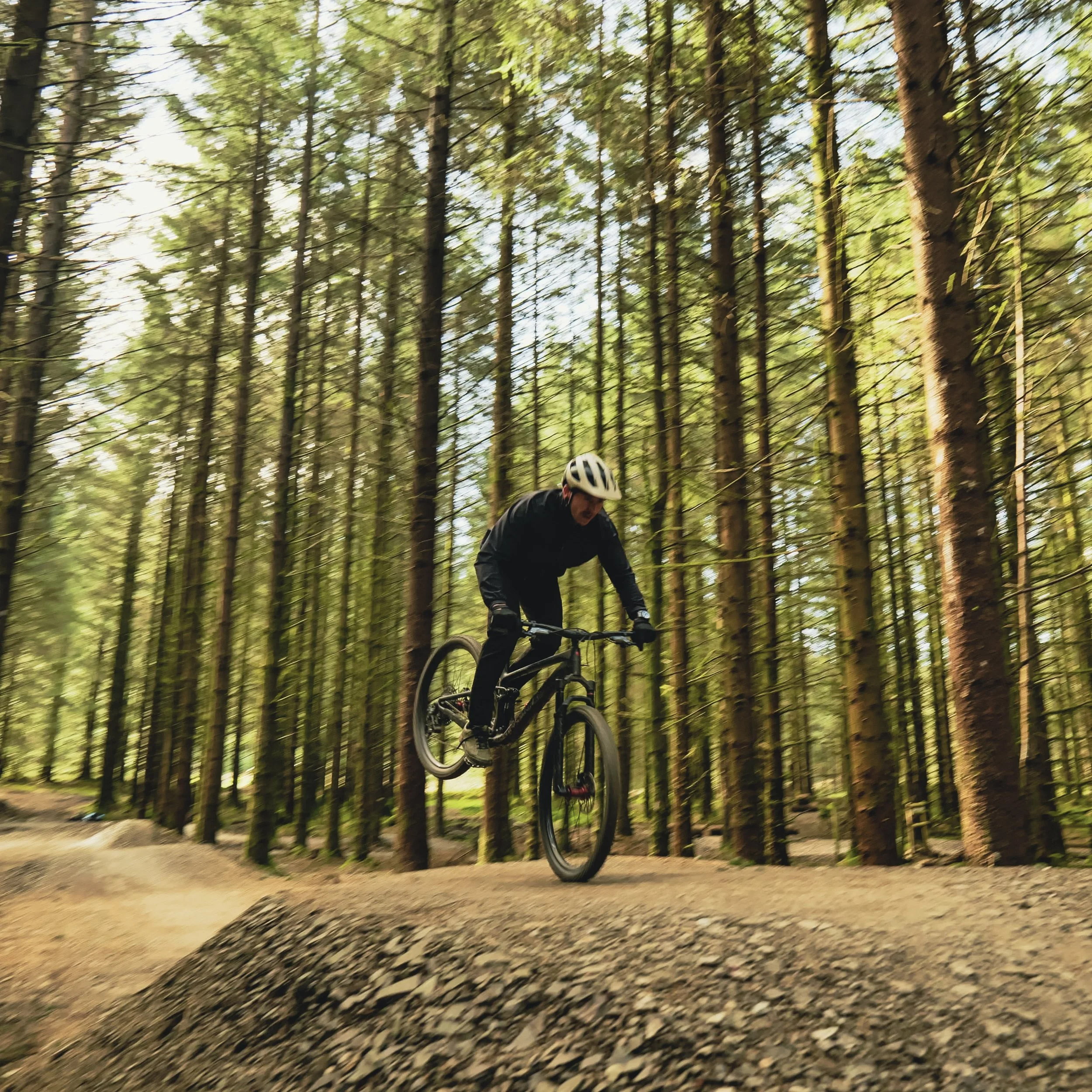 A person riding a mountain bike in a forest, mid-air over a dirt trail obstacle, wearing a helmet and dark clothing.