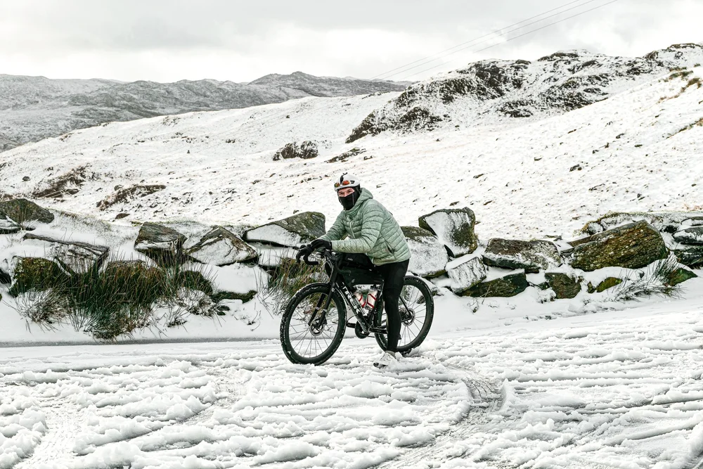Daniil wheeling his bike along a snowy mountain road beside a stone wall