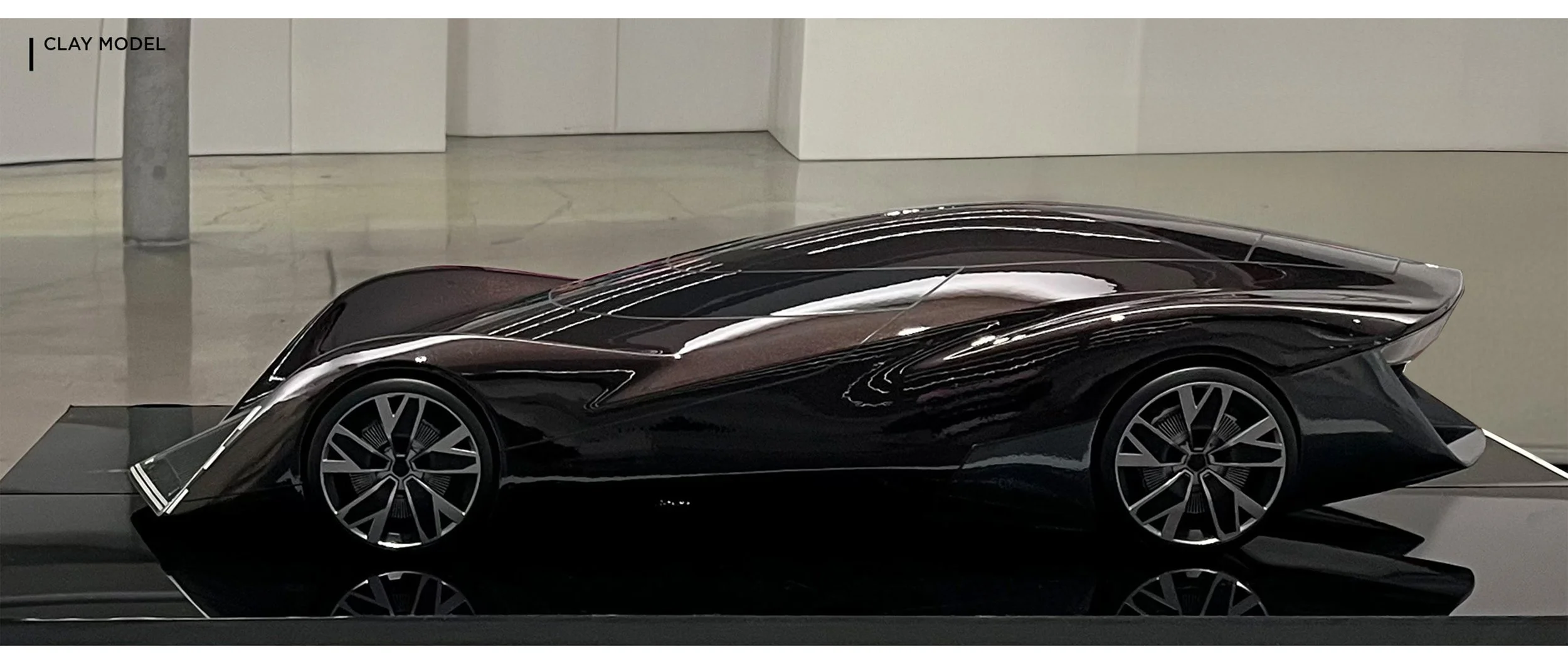 A sleek, futuristic black and brown clay model of a concept sports car on display indoors. The car has sharp, aerodynamic lines and distinctive rims.