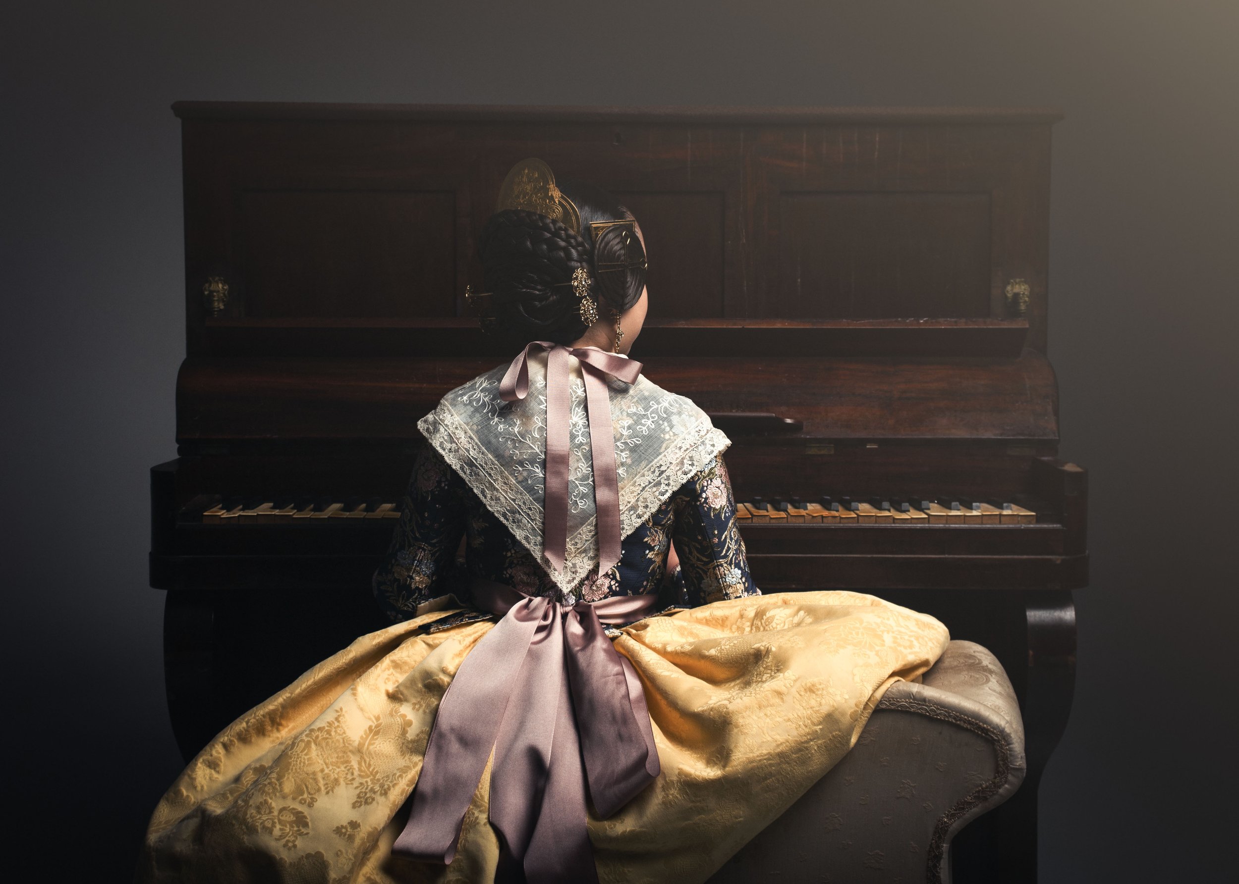 Mujer vestida con ropa tradicional fallera sentada frente a un piano de madera, vista de espaldas, en un entorno de luz suave y ambiente clásico.