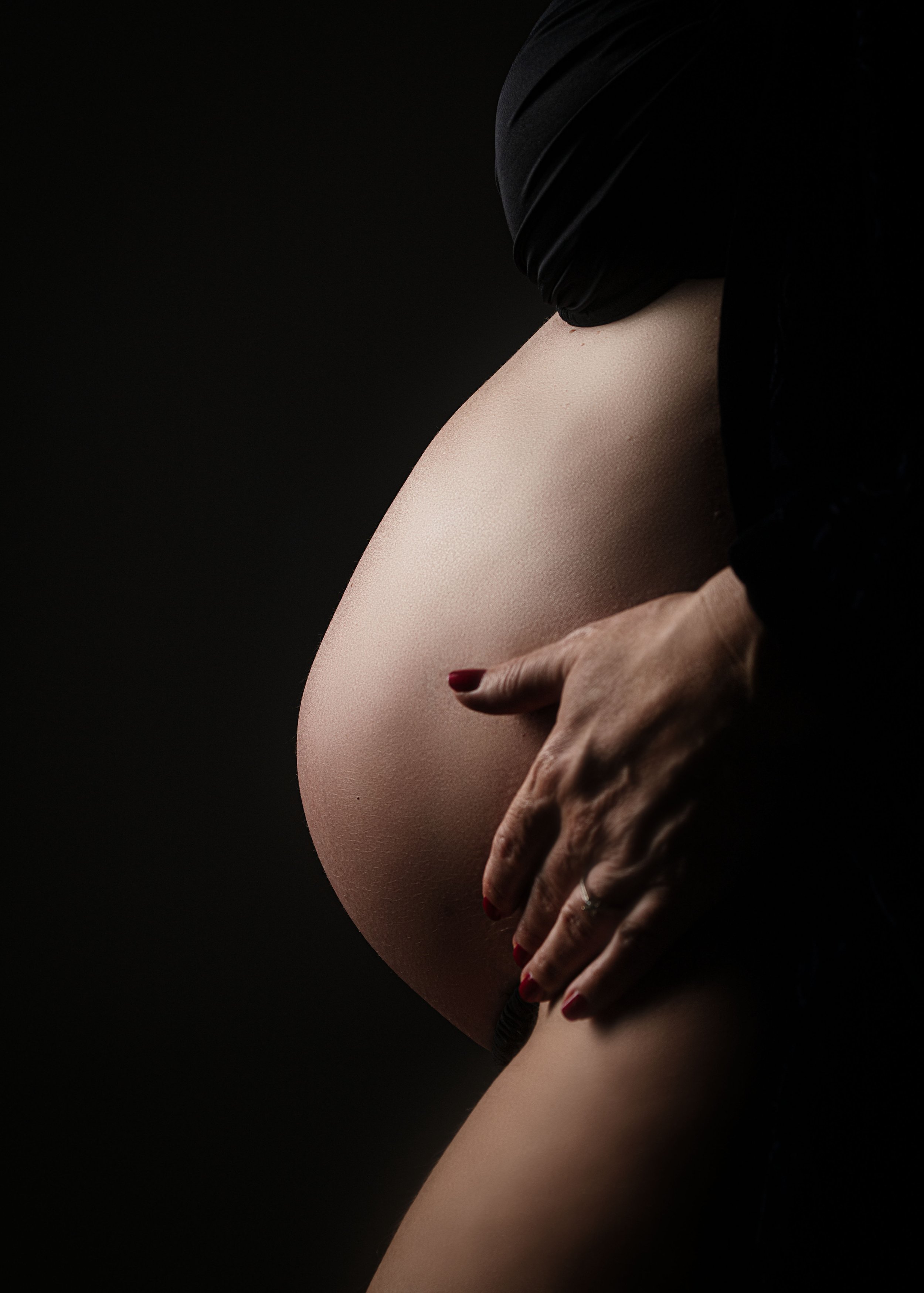 Pierna y cadera de una mujer embarazada , tocándose la barriga  suavemente, con fondo negro y ropa oscura.