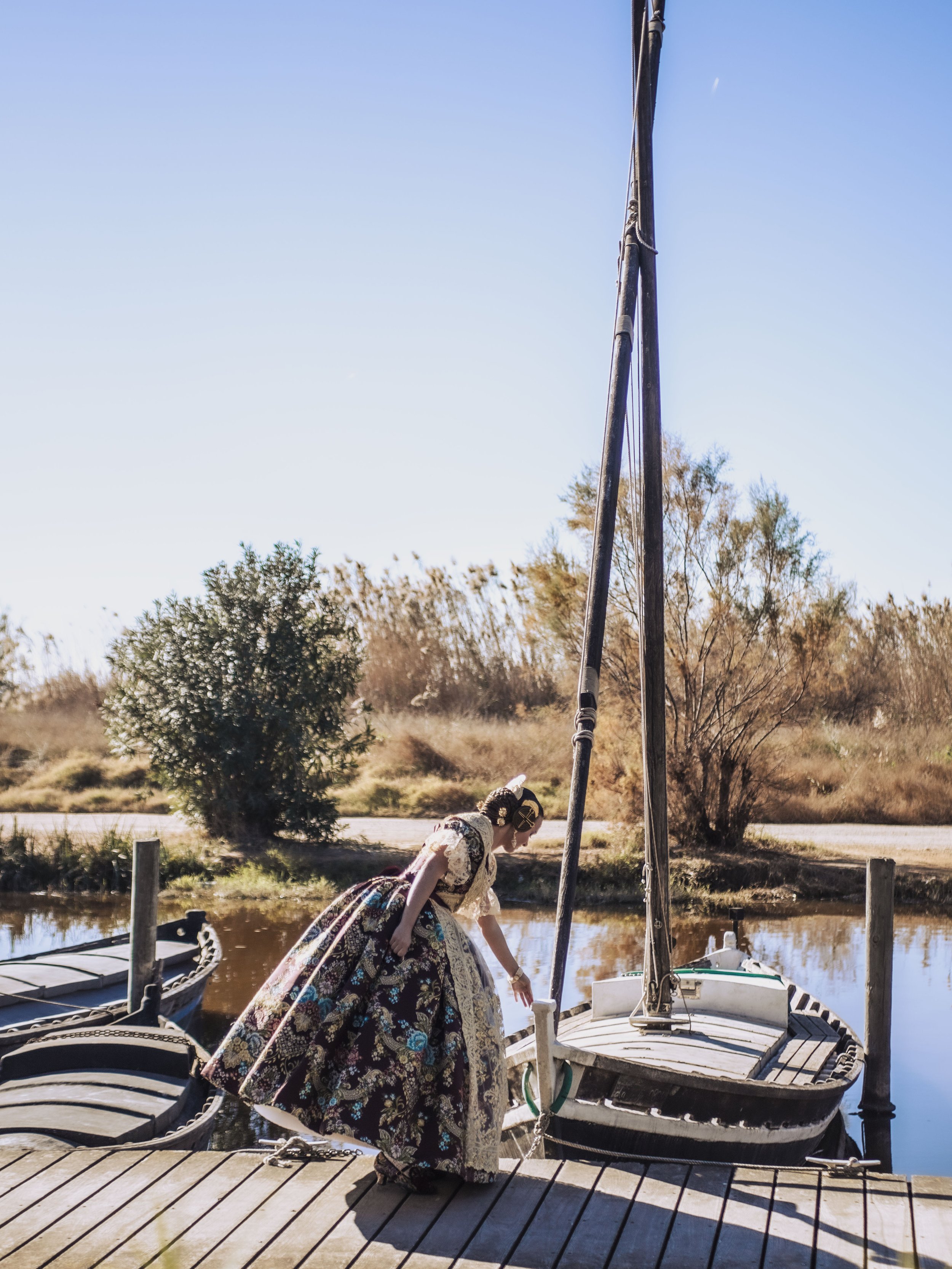 Mujer vestida con traje tradicional en un muelle, junto a un barco de vela en un paisaje natural con árboles y agua.