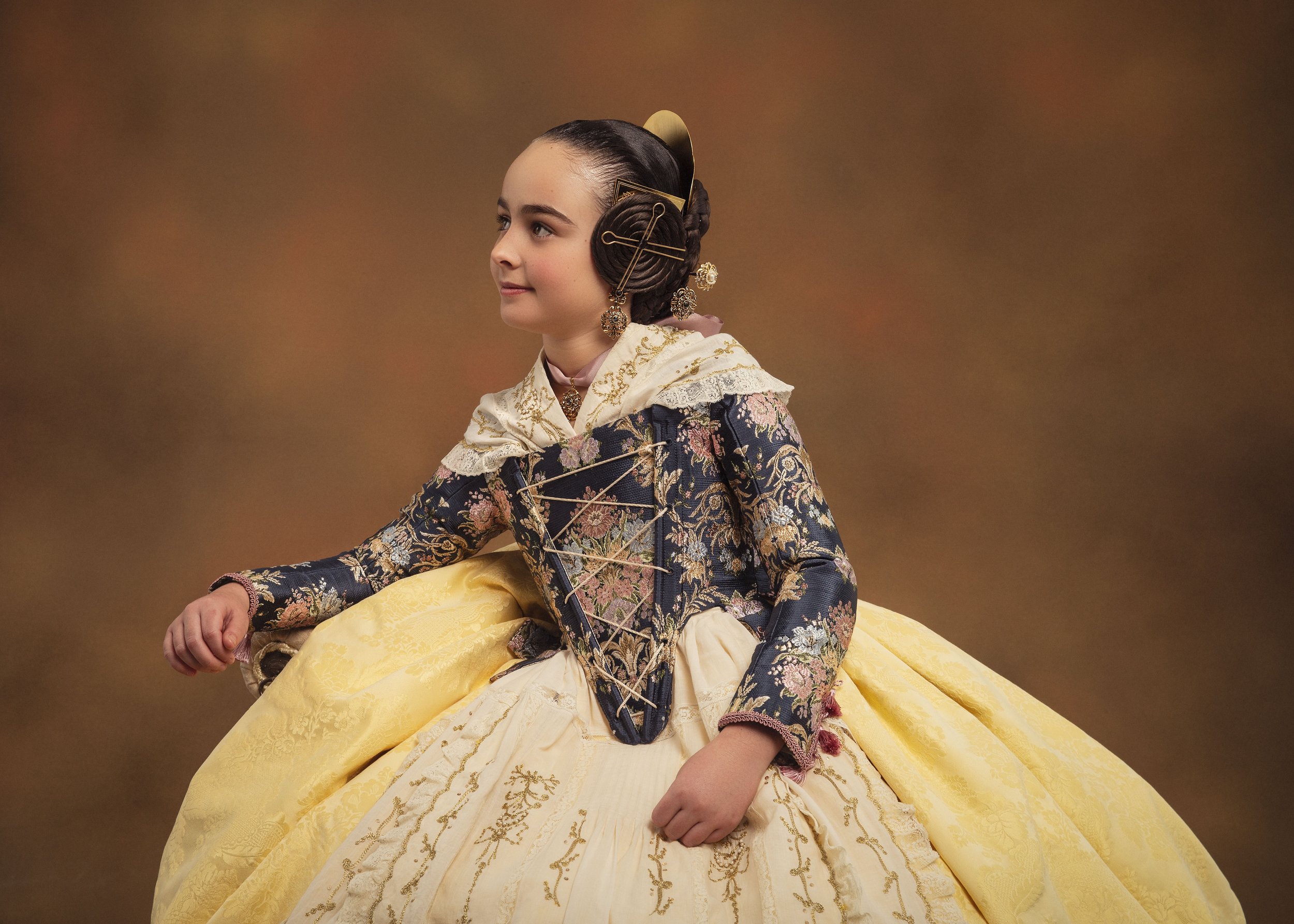 Niña vestida con traje de época de color amarillo y una chaqueta con detalles florales, peinada con accesorios tradicionales y un fondo marrón