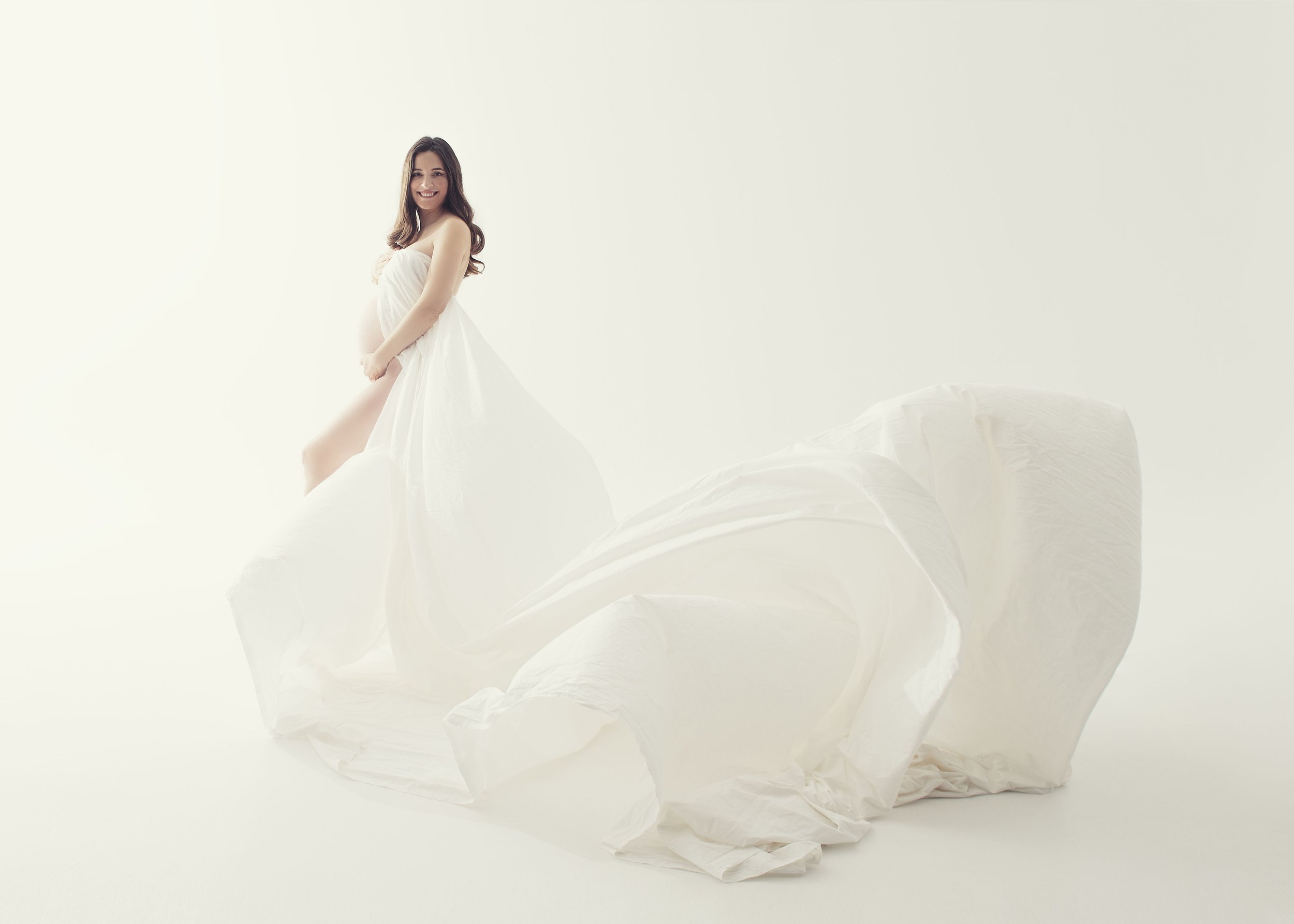 Mujer embarazada en vestido blanco, sonriendo, sentada sobre una tela blanca en un fondo claro.