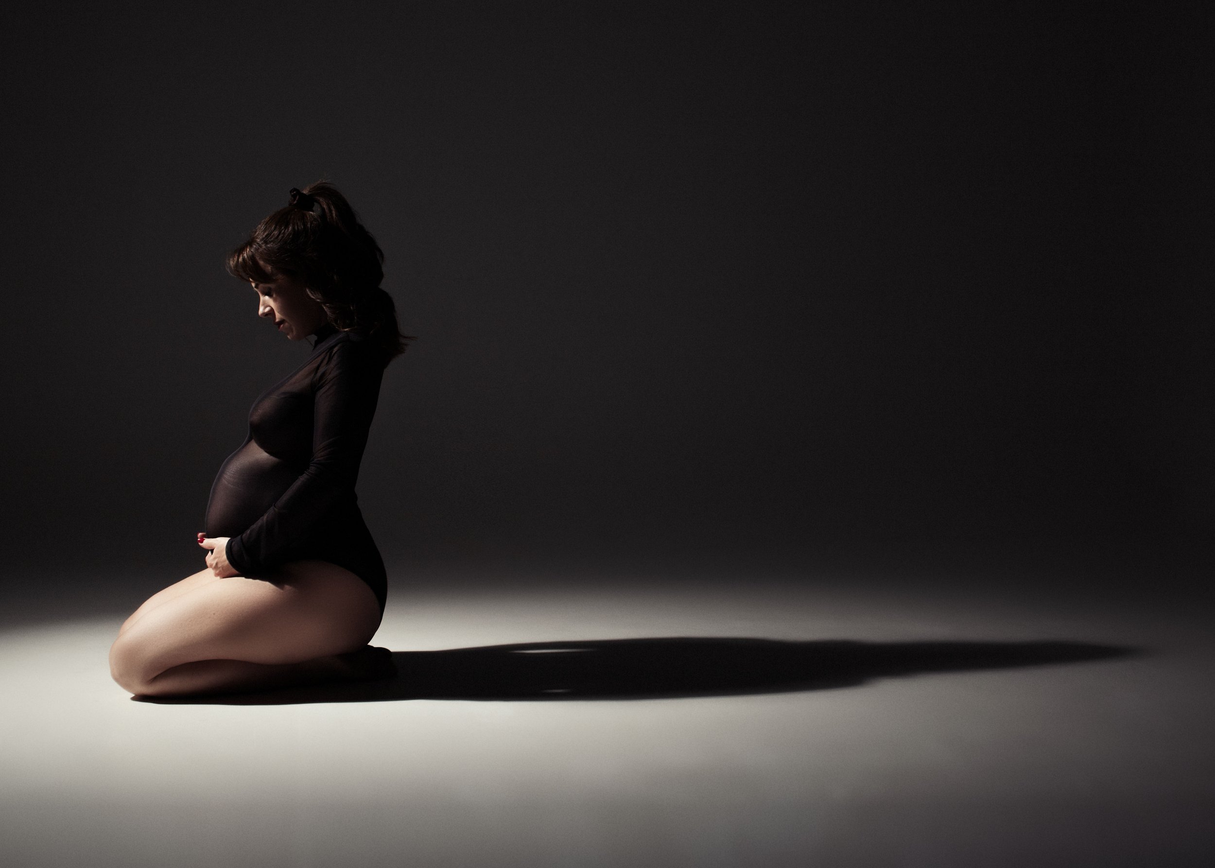 Mujer embarazada arrodillada  en un fondo oscuro.