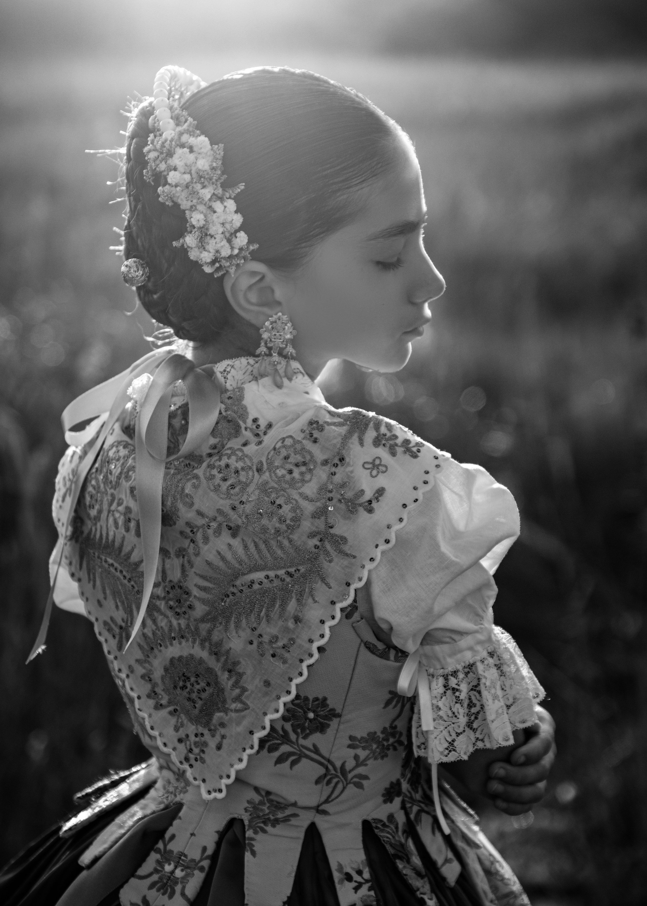 Retrato en blanco y negro de una joven con ojos cerrados, vestida con ropa tradicional valenciana , con peinado adornado con flores y accesorios, en un entorno natural.