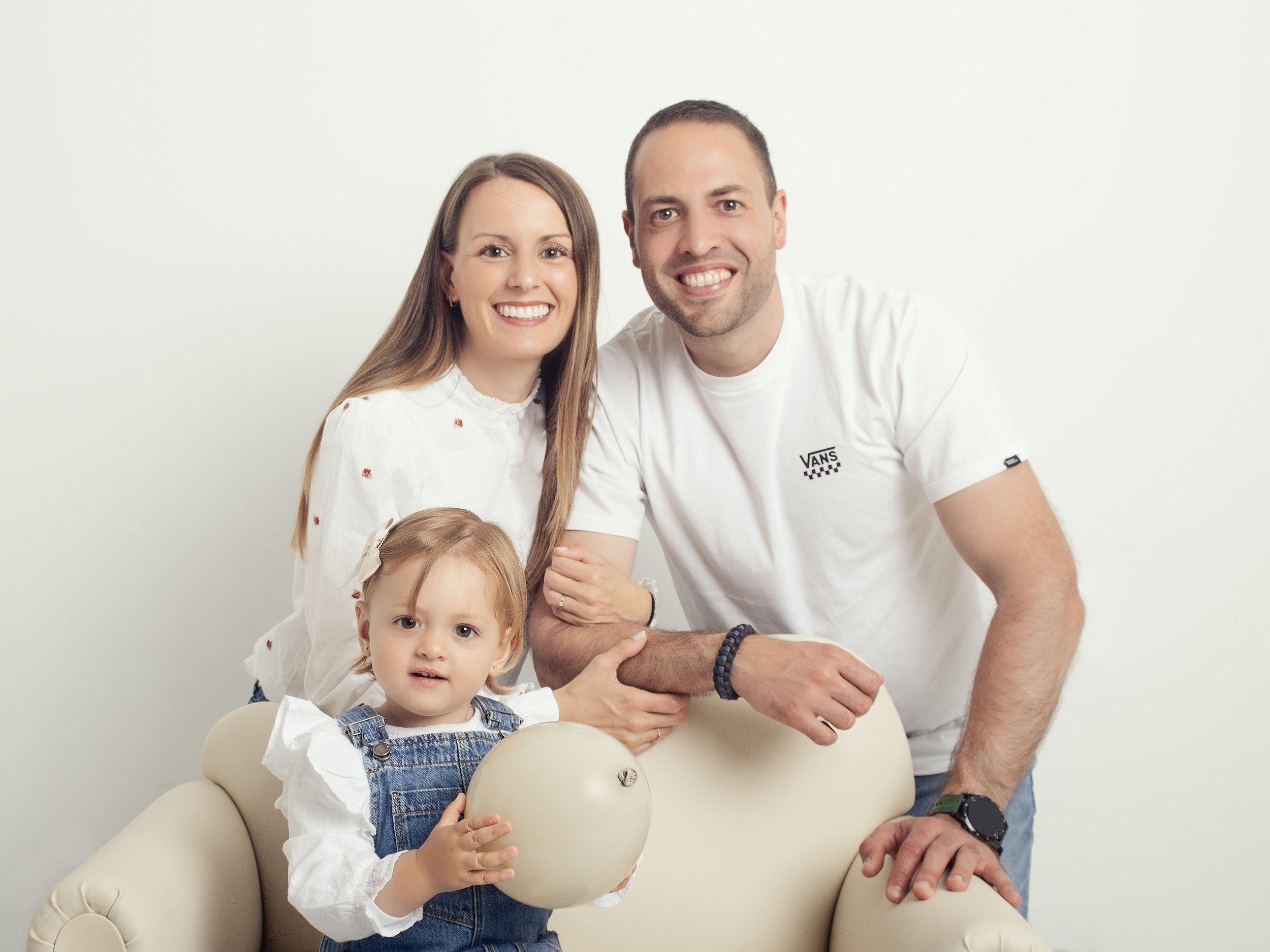 Familia joven sonriendo en una sesión de fotos en interior, con una niña pequeña sosteniendo un globo beige.