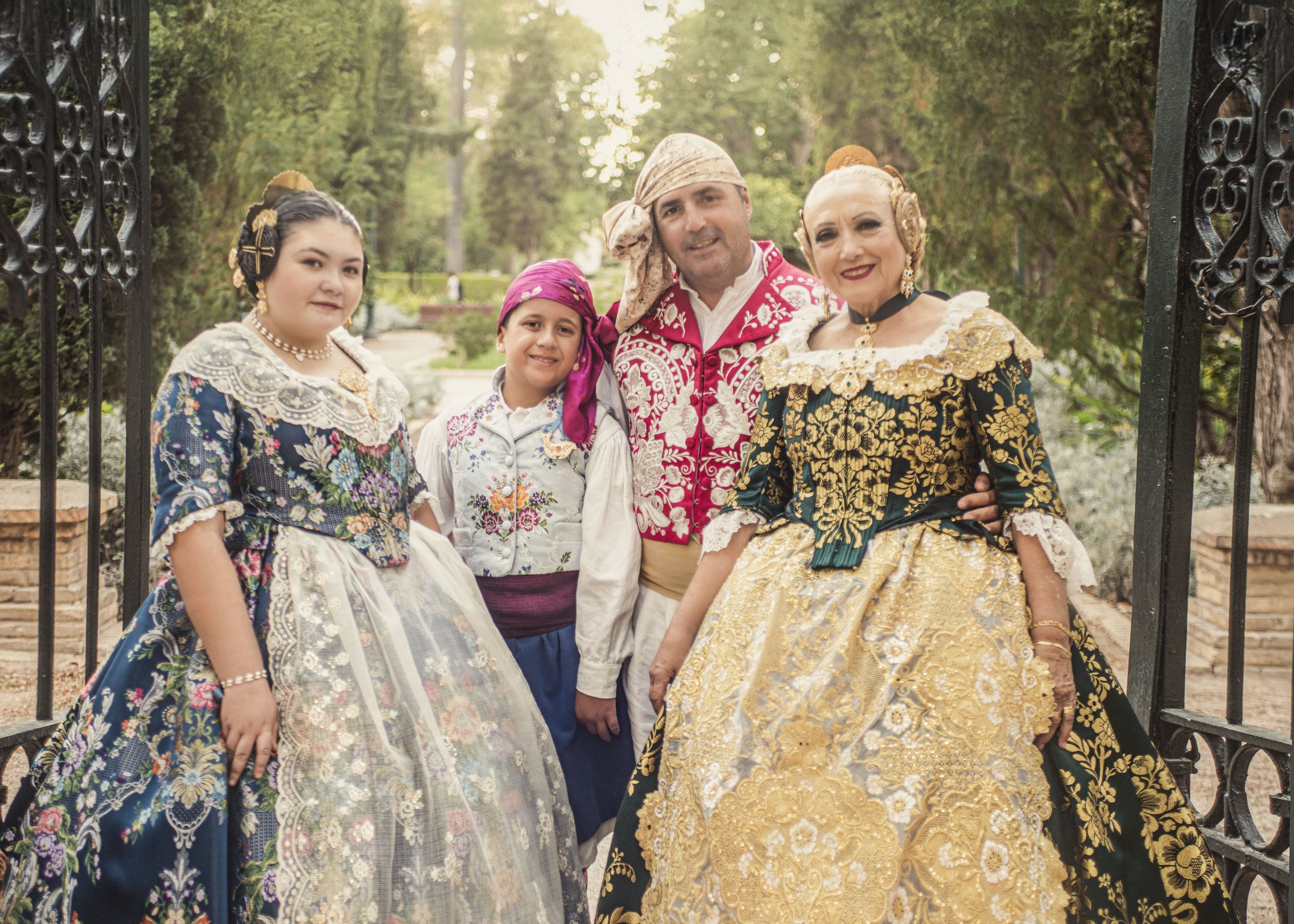 Cuatro personas vestidas con trajes tradicionales valencianos posando en un parque con árboles y una entrada de hierro en el fondo.