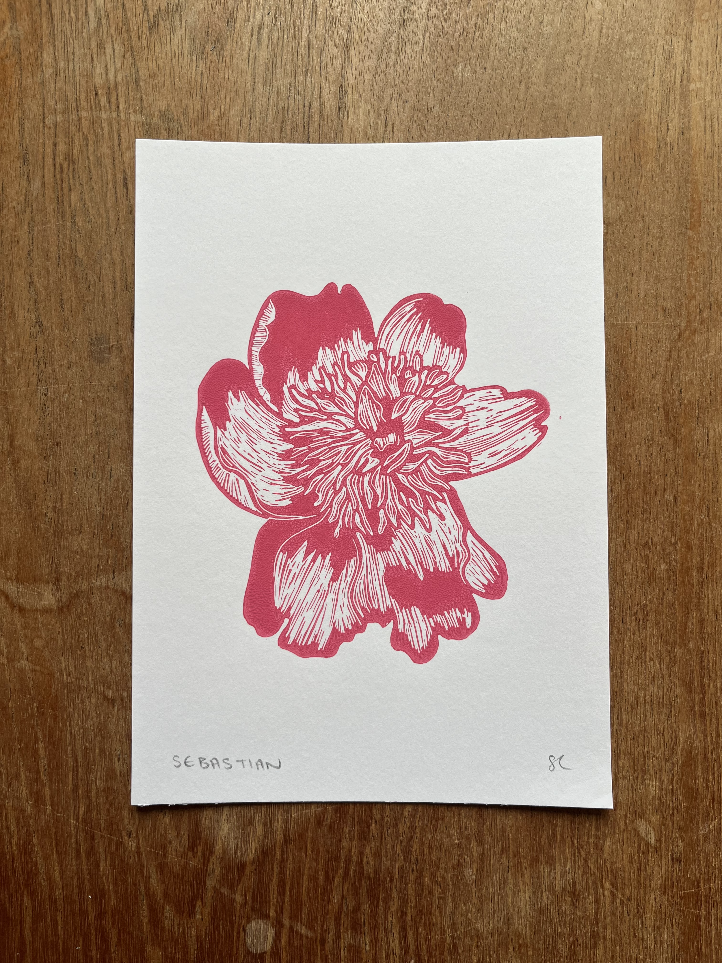 A white paper with a pink line art illustration of a flower, with the name 'SEBASTIAN' handwritten in the bottom left corner and initials 'SC' in the bottom right corner, placed on a wooden surface.