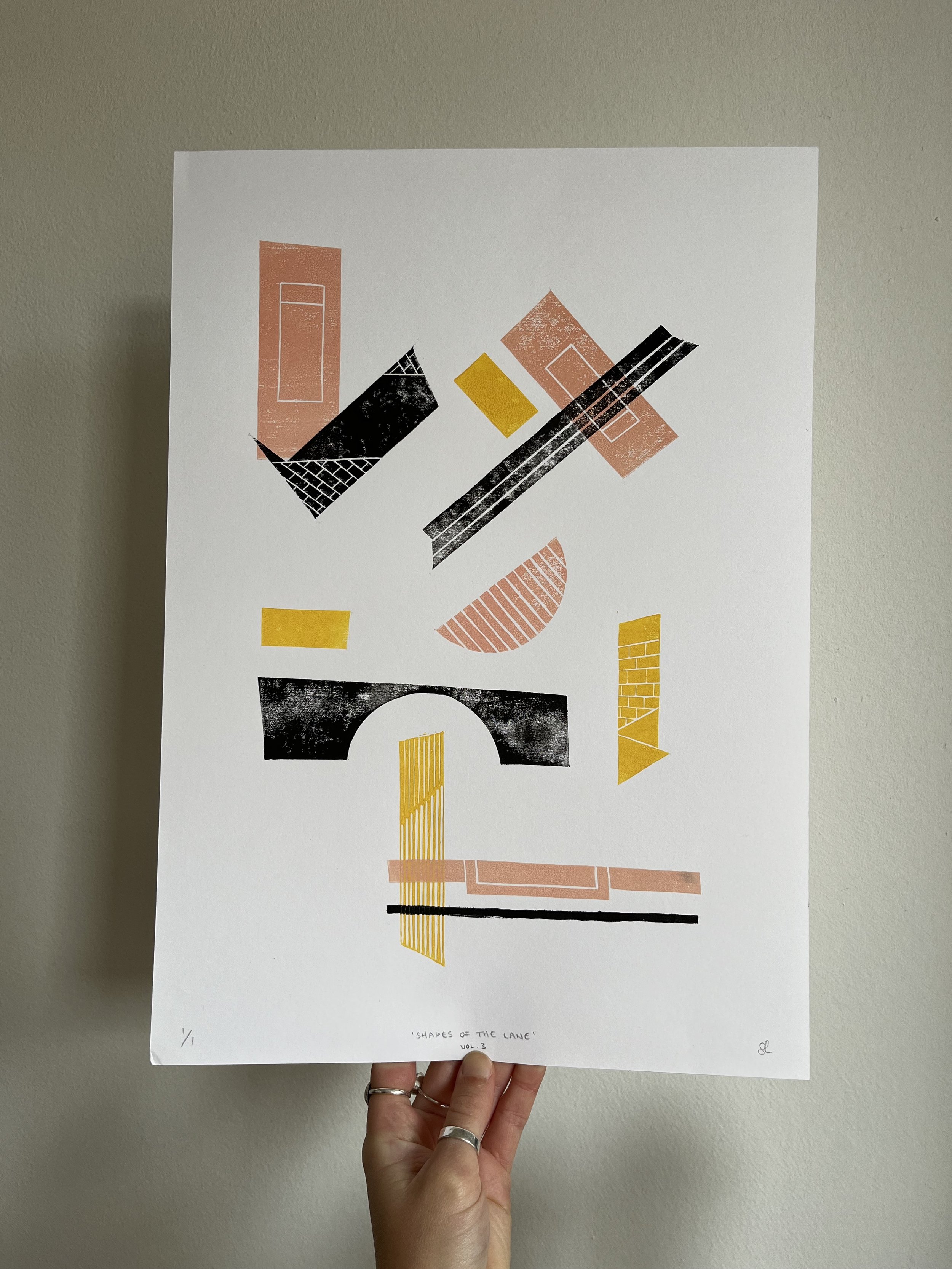 An abstract art print titled 'Shapes of the Lane Vol. 3' with various geometric shapes in pink, black, yellow, and orange on white paper held against a beige wall.
