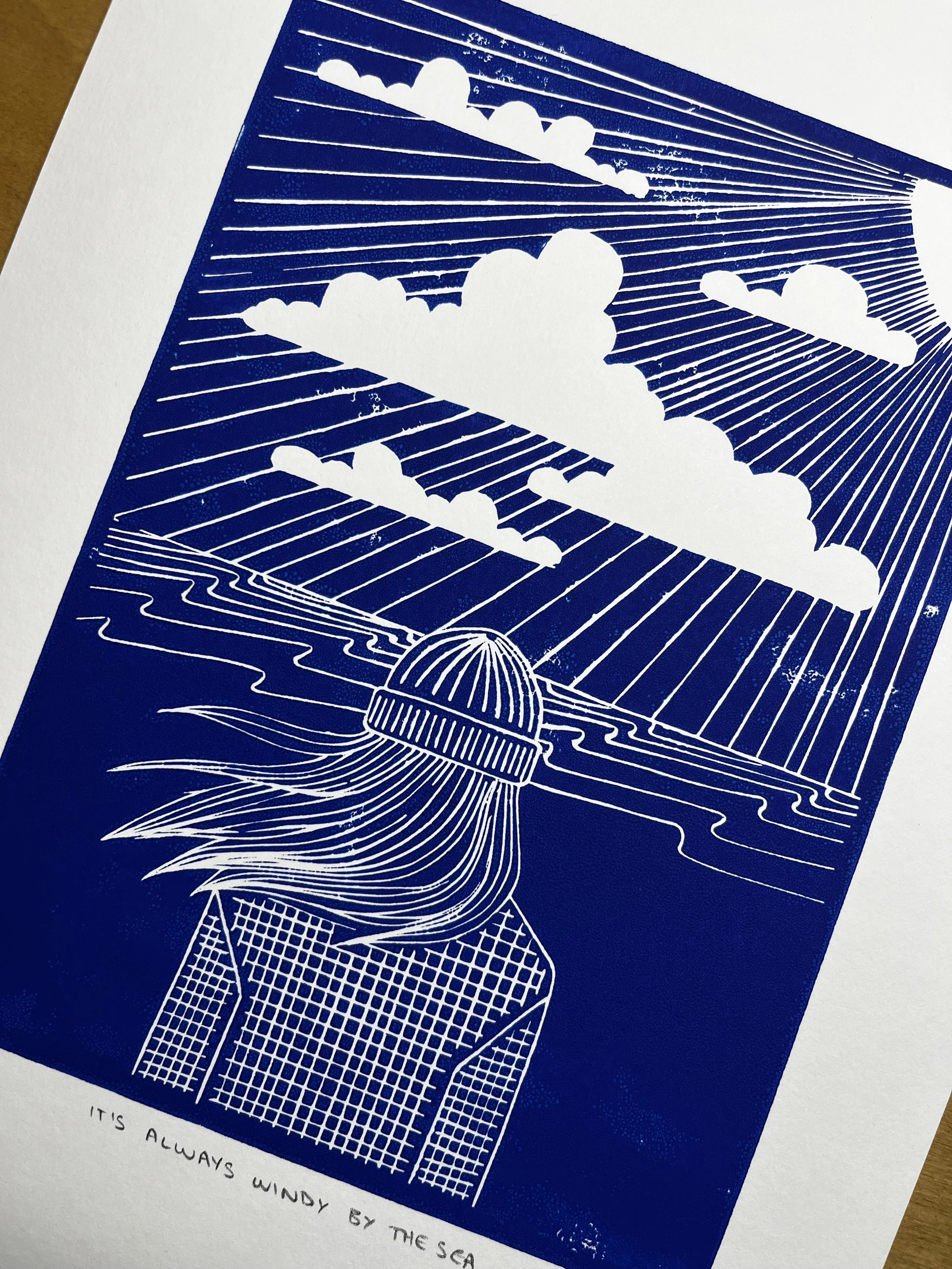 A line art illustration of a girl with flowing hair wearing a hat, looking at clouds and the sea, with the sun's rays shining down. Text at bottom reads 'It's always windy by the sea.'
