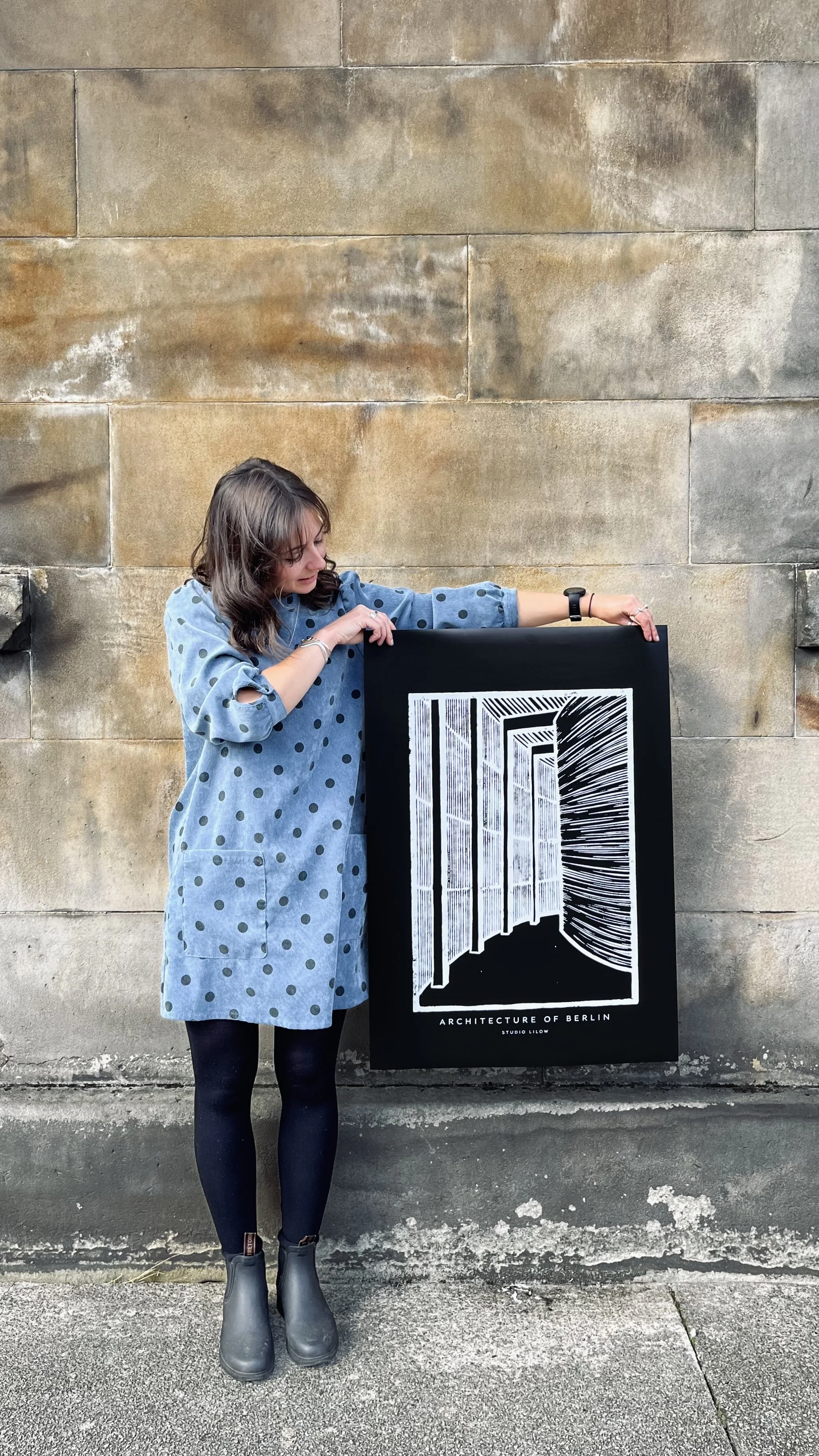 A woman wearing a blue polka dot dress and black tights holding a black poster board with a white barcode-style graphic and architectural lines, standing against a stone wall.
