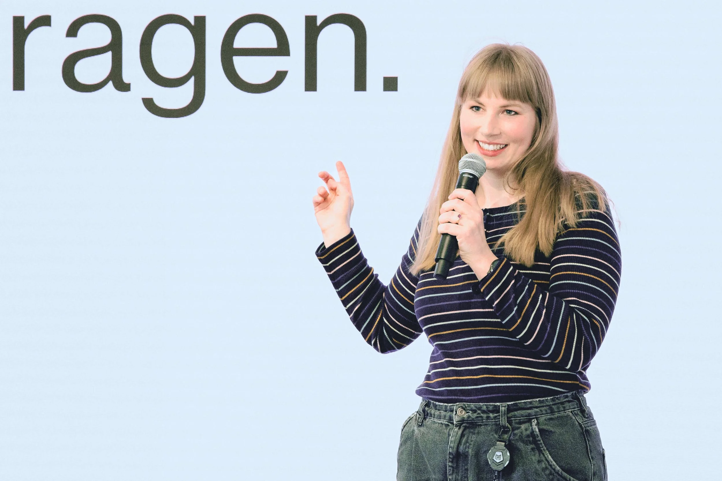 Eine junge Frau mit langen blonden Haaren hält ein Mikrofon in der Hand und spricht vor einer hellblauen Wand mit schwarzer Schrift.