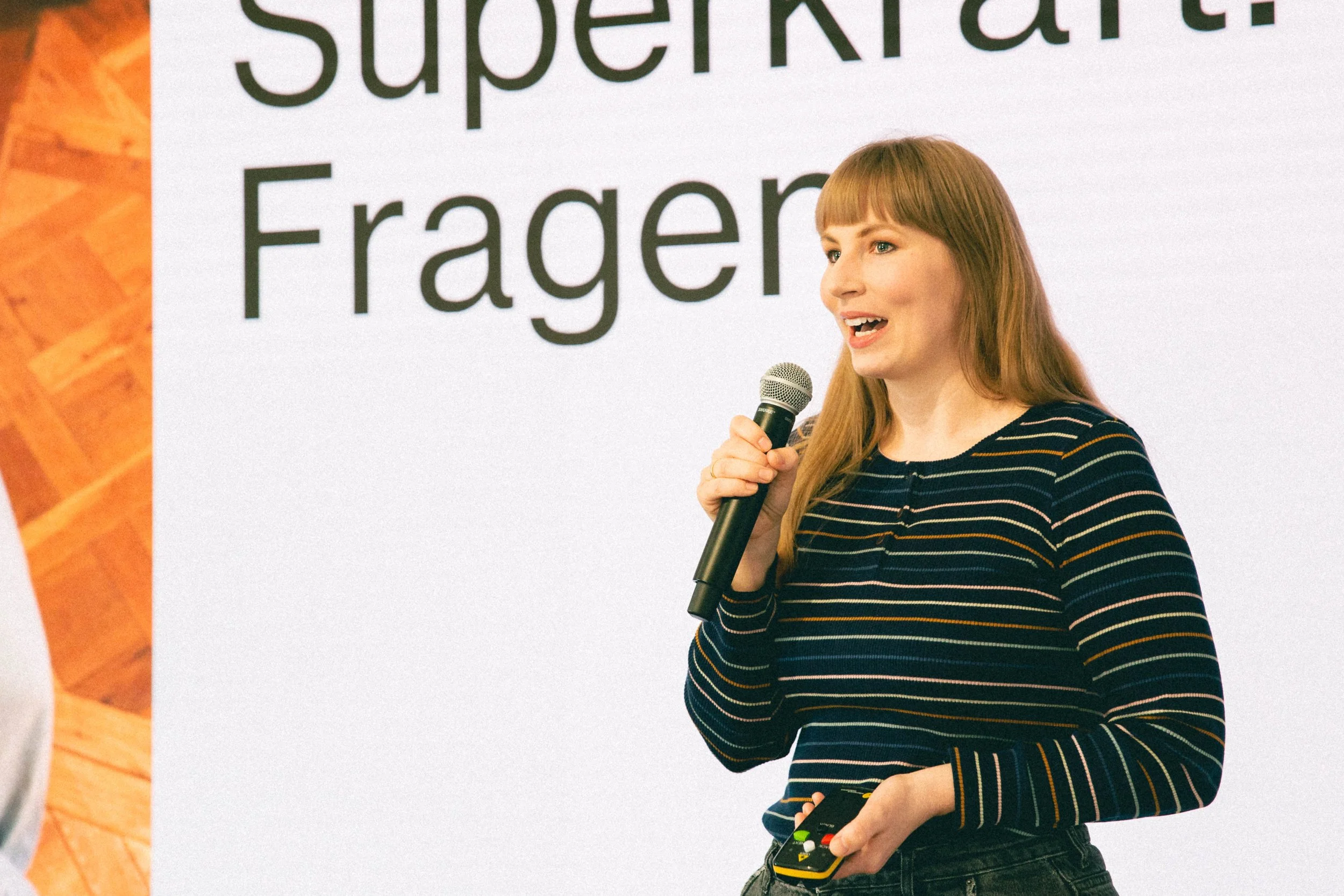 Eine junge Frau hält ein Mikrofon in der Hand und spricht vor einer großen Präsentationsfolie, auf der das Wort 'Fragen' in deutscher Sprache steht.