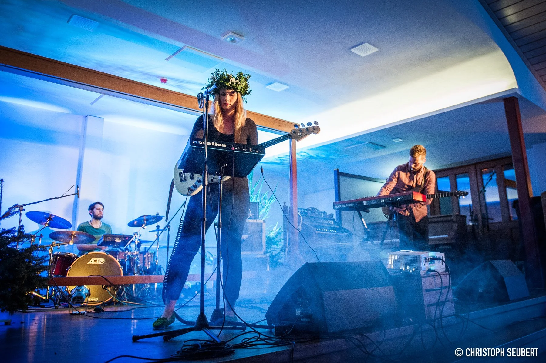 Eine Band mit drei Mitgliedern spielt auf einer Bühne: eine weibliche Sängerin mit einer Gitarre, ein männlicher Keyboarder und ein männlicher Schlagzeuger im Hintergrund. Es gibt blaue Beleuchtung und Nebel auf der Bühne.