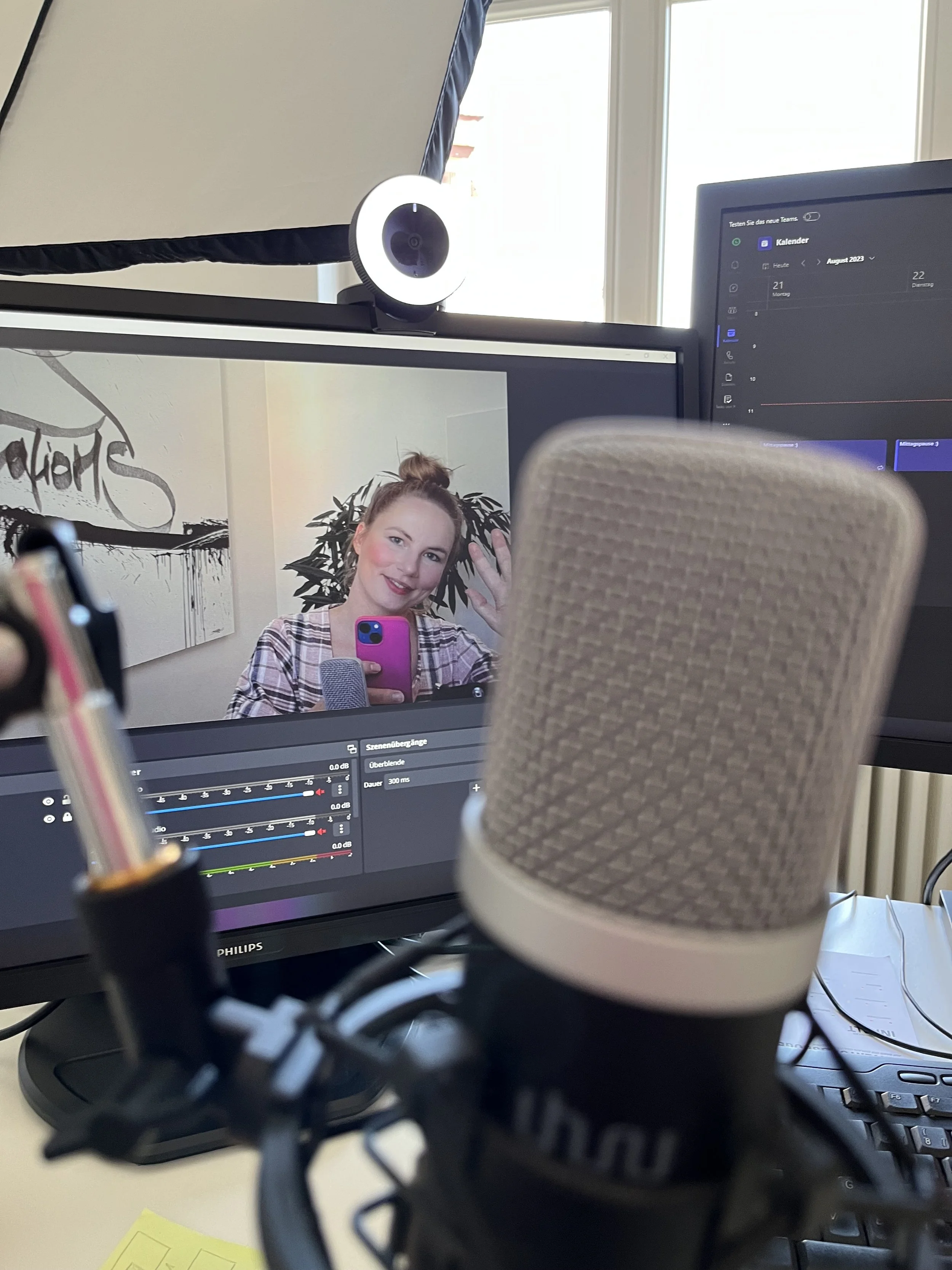 Ein Aufnahmegerät, ein Mikrofon und zwei Monitore sind auf einem Schreibtisch zu sehen. Auf dem Bildschirm ist eine Frau zu sehen, die in die Kamera lächelt und ein Selfie macht.