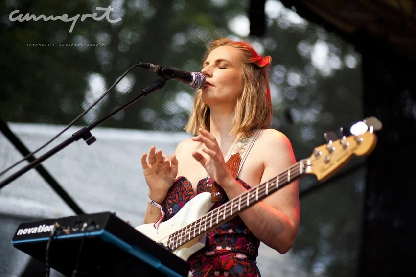 Eine junge Frau mit einem E-Bass, die singt und auf einer Bühne im Freien spielt.