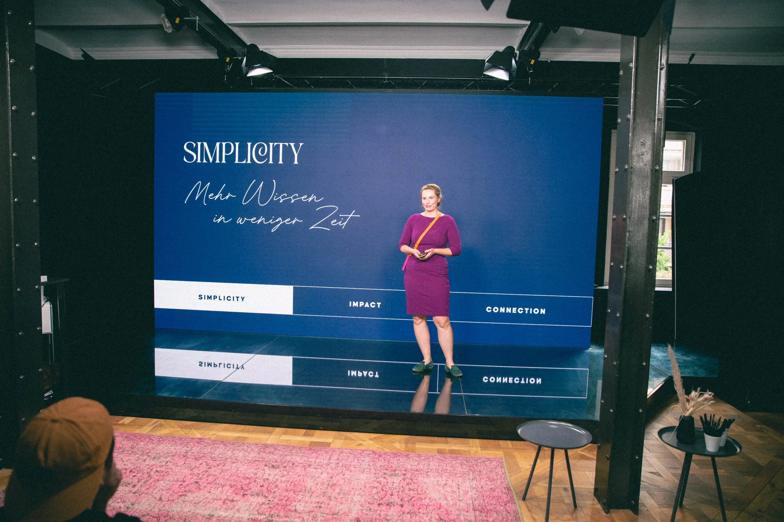 Eine Frau steht vor einem großen Bildschirm, auf dem steht: 'SIMPLICITY Mehr Wissen in weniger Zeit' und die Wörter 'Simplicity', 'Impact' und 'Connection' sind auf einer Balken-Diagrammlinie sichtbar.