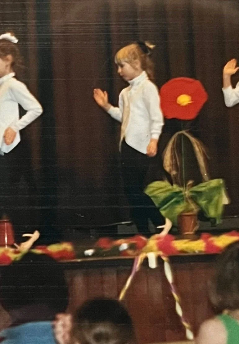 Kinder auf einer Bühne bei einer Schulaufführung, einige Kinder in weißen Hemden, ein Kind trägt eine rote Mütze, dekorative Blumen und Pflanzen sind auf der Bühne.