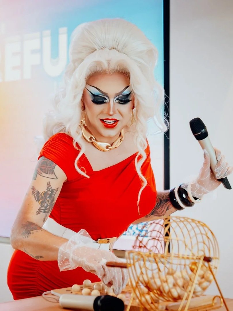 Une drag queen aux cheveux blonds bouclés, maquillage extravagant, portant une robe rouge, tenant un micro et manipulant une machine à boule à neige.