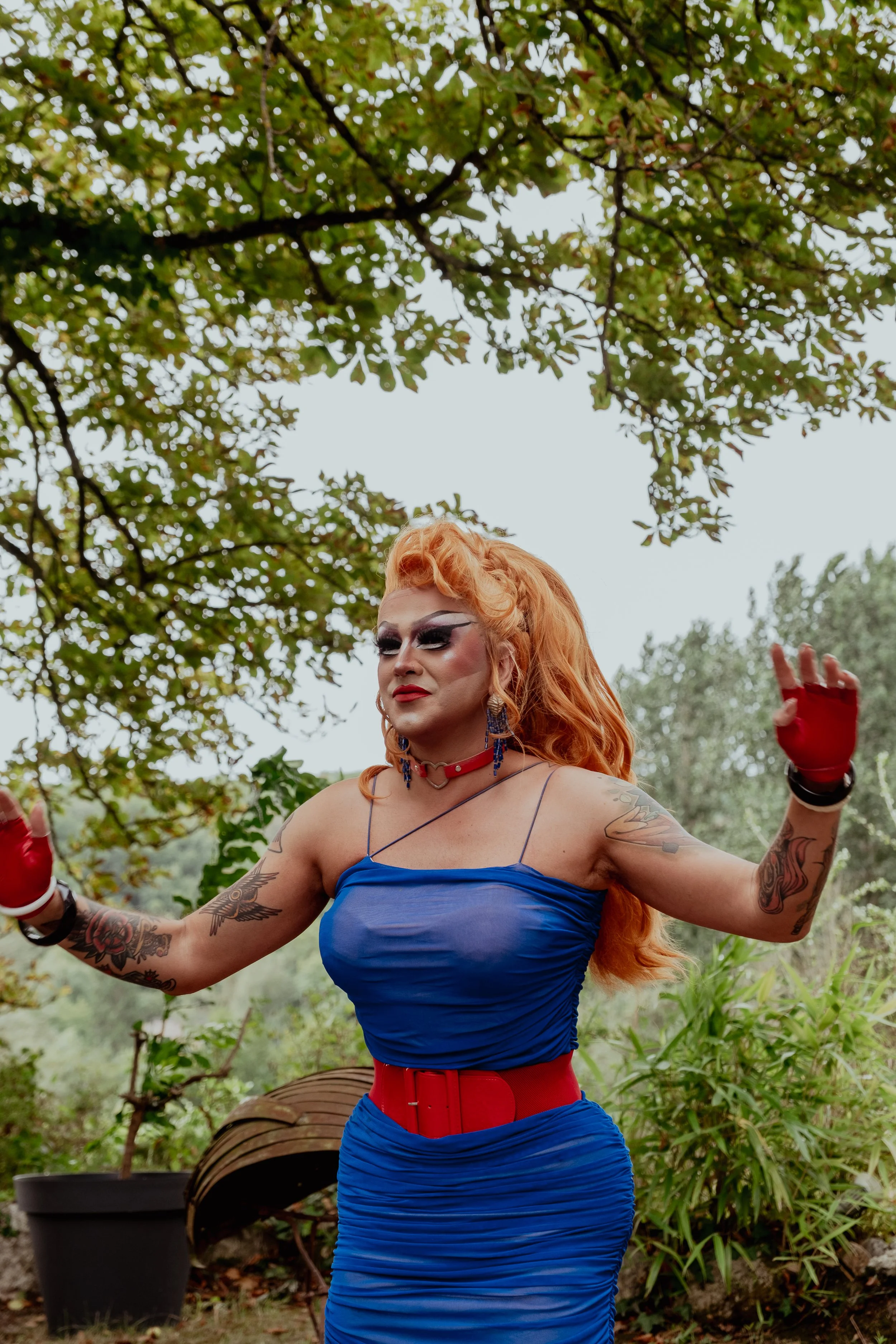 Une personne avec des tatouages, portant une robe bleue et un bel ensemble de maquillage, se tient dans un environnement naturel, avec des arbres en arrière-plan.