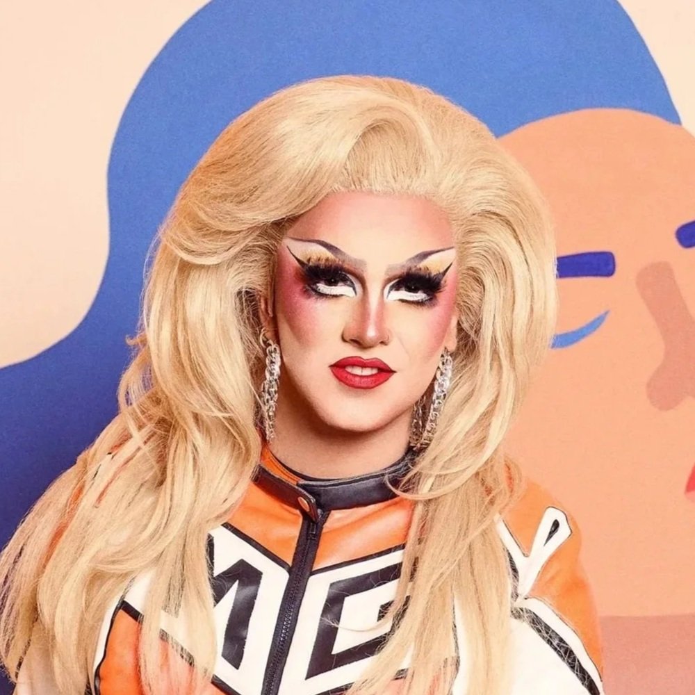 Une personne avec un maquillage coloré et une perruque blonde, portant des boucles d'oreilles longues, pose devant un fond coloré avec un visage stylisé.